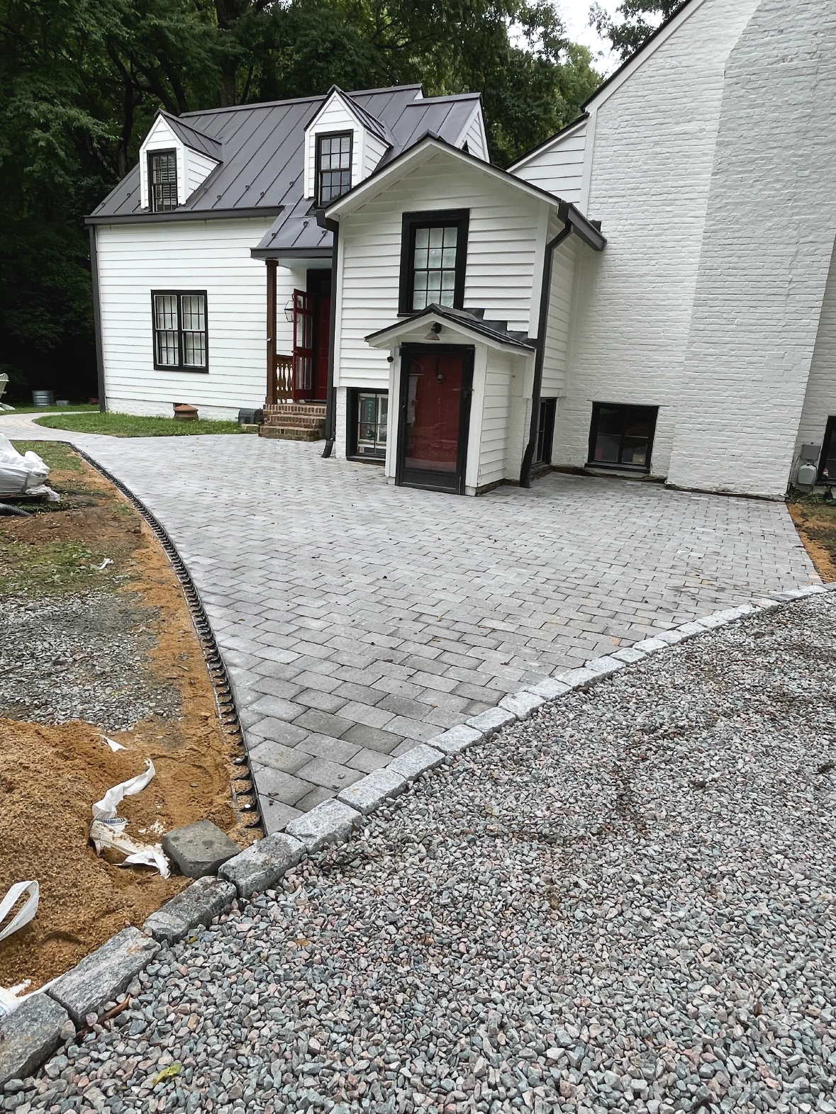 Newly paved driveway and walkway with gray bricks, white house with black window frames, landscaped area with gravel and dirt, surrounded by greenery.