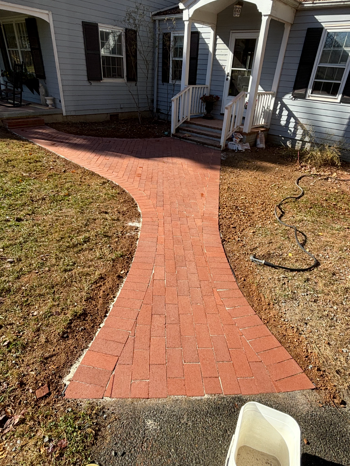 A newly paved brick pathway leading to the front porch of a house with blue siding and white trim. The yard around the path is partly dug up, with soil and a garden hose on the ground.