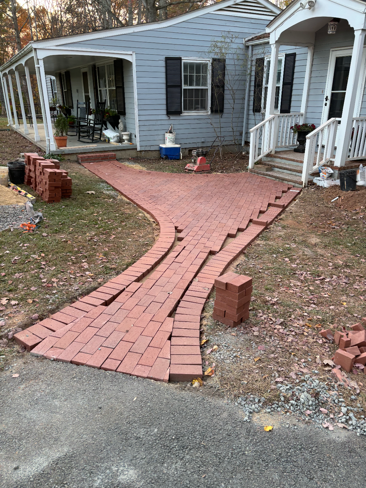 Brick pathway under construction leading to the front porch of a house, with some stacked bricks and construction tools nearby.