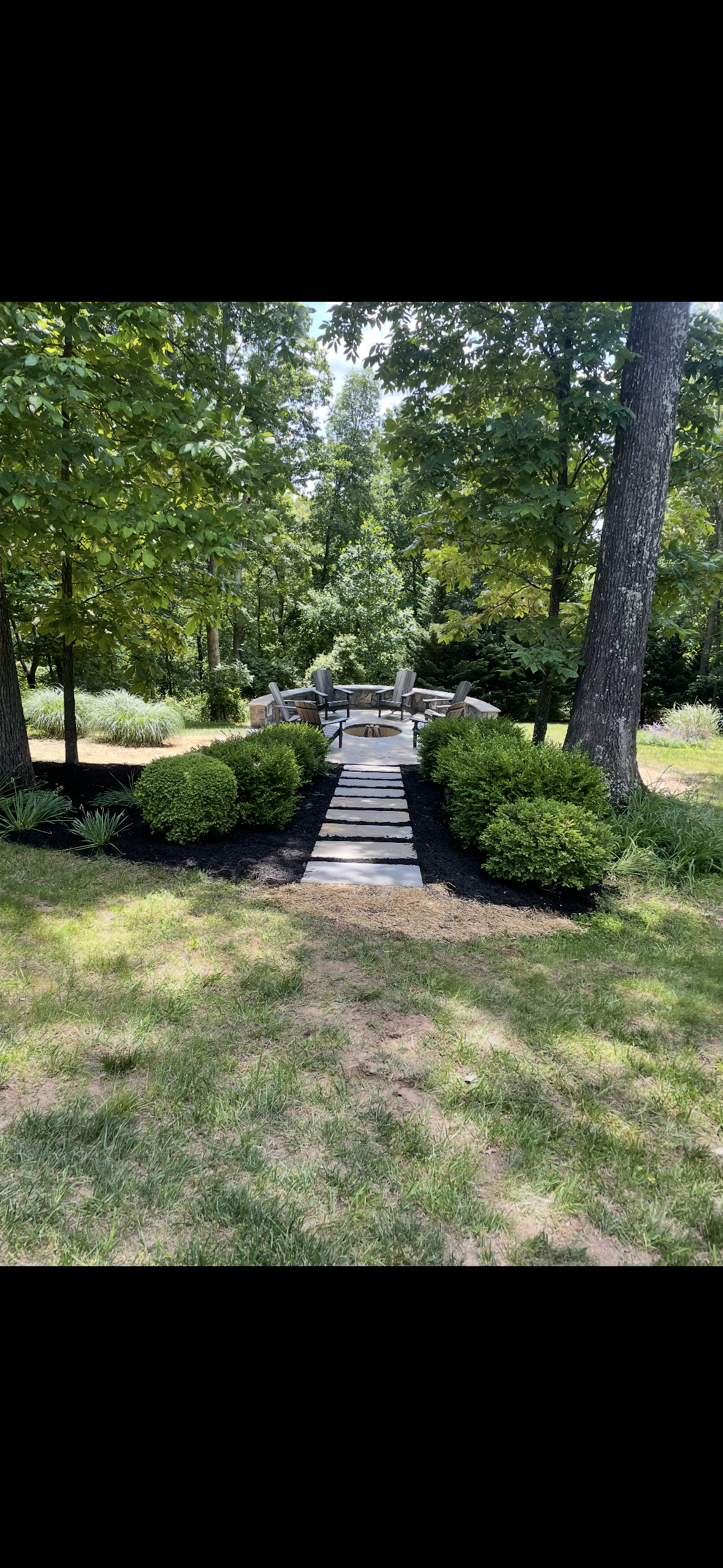 A landscaped backyard with a stone pathway leading to a circular fire pit surrounded by Adirondack chairs. Green shrubs and tall trees frame the scene on a sunny day.