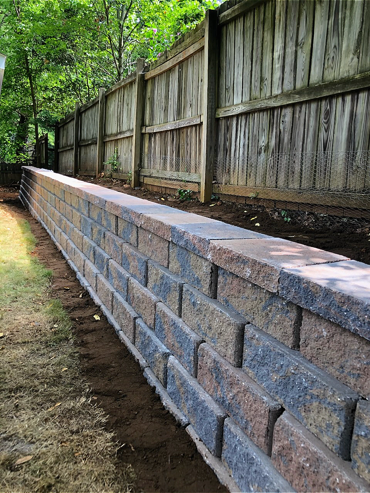 A newly built stone retaining wall along a grassy yard, with a wooden fence in the background and trees overhead.