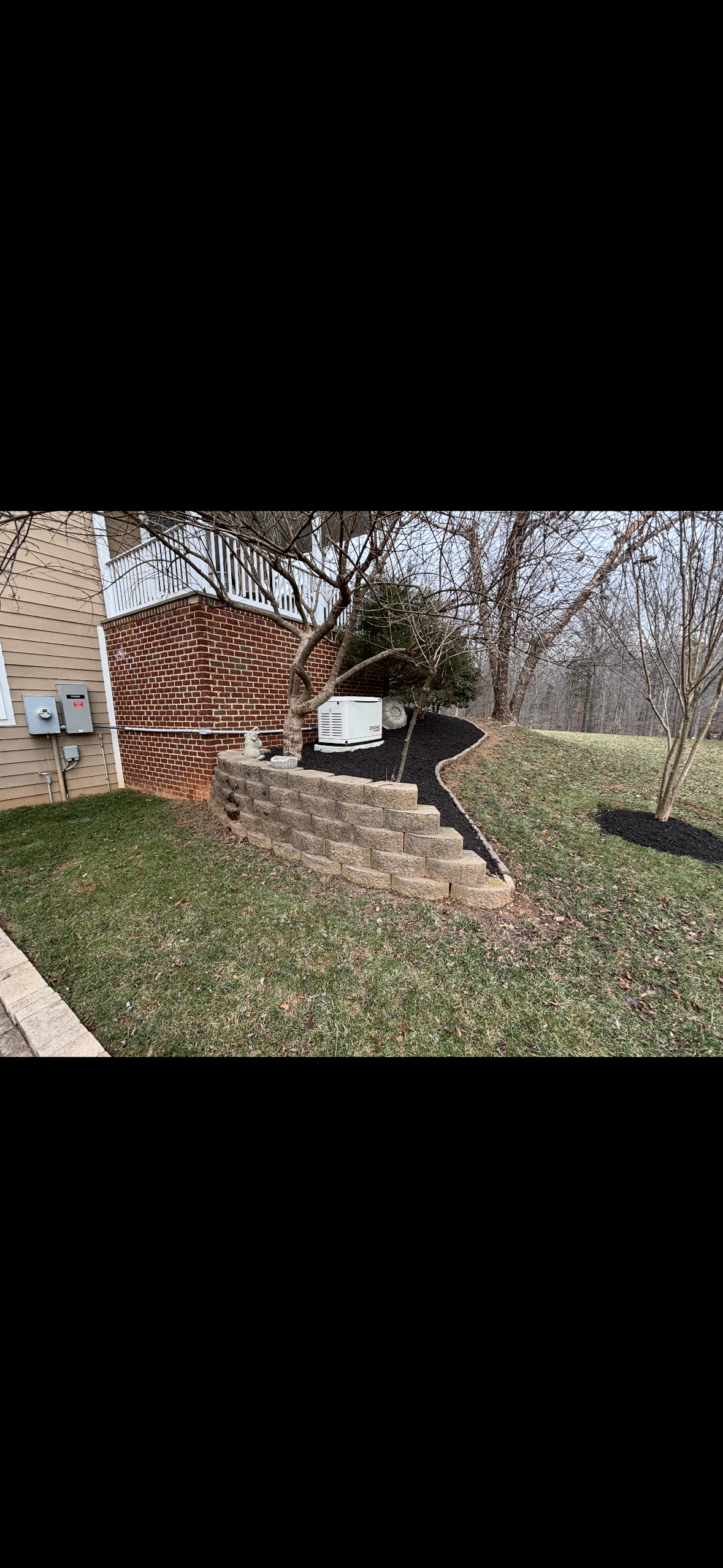 A backyard with a brick house wall, leafless trees, a curved stone retaining wall, a small set of steps made from stone blocks, and black mulch around the plants and trees.