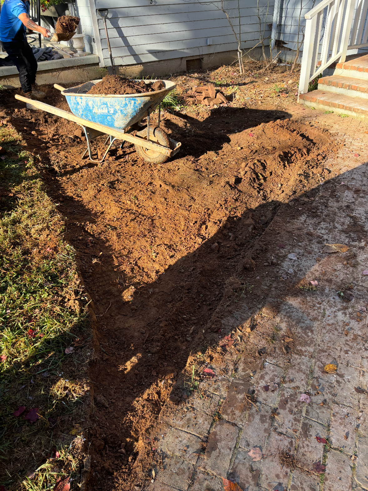 A person planting or preparing a garden bed outside of a house, using a wheelbarrow filled with soil and shoveling dirt onto the ground.