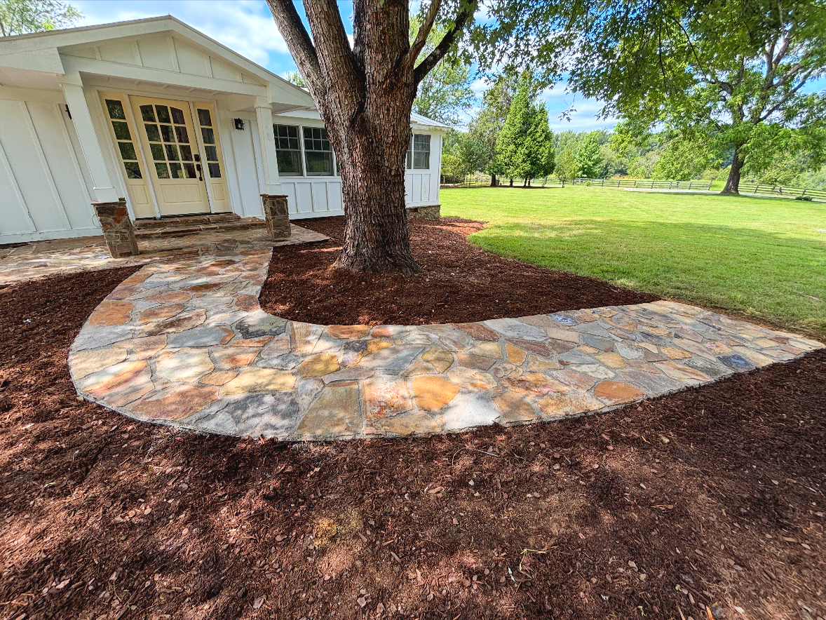 A curved stone walkway leads from a house to a grassy yard, with a tree in the center surrounded by mulch. The house is white with a porch and multiple windows.