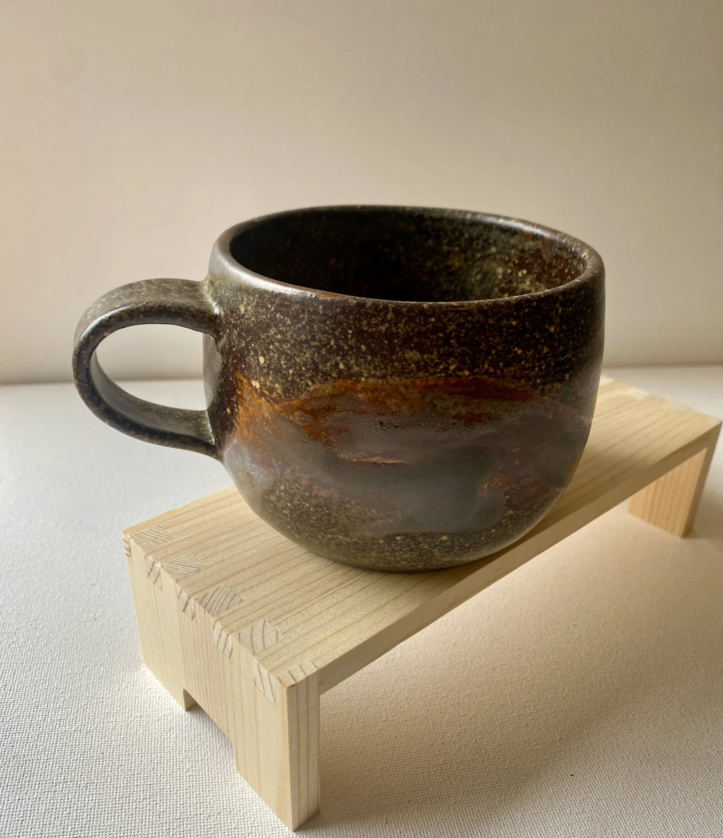view 1 of brown mug with splatter and metallic designs in the glaze sitting on wood stand