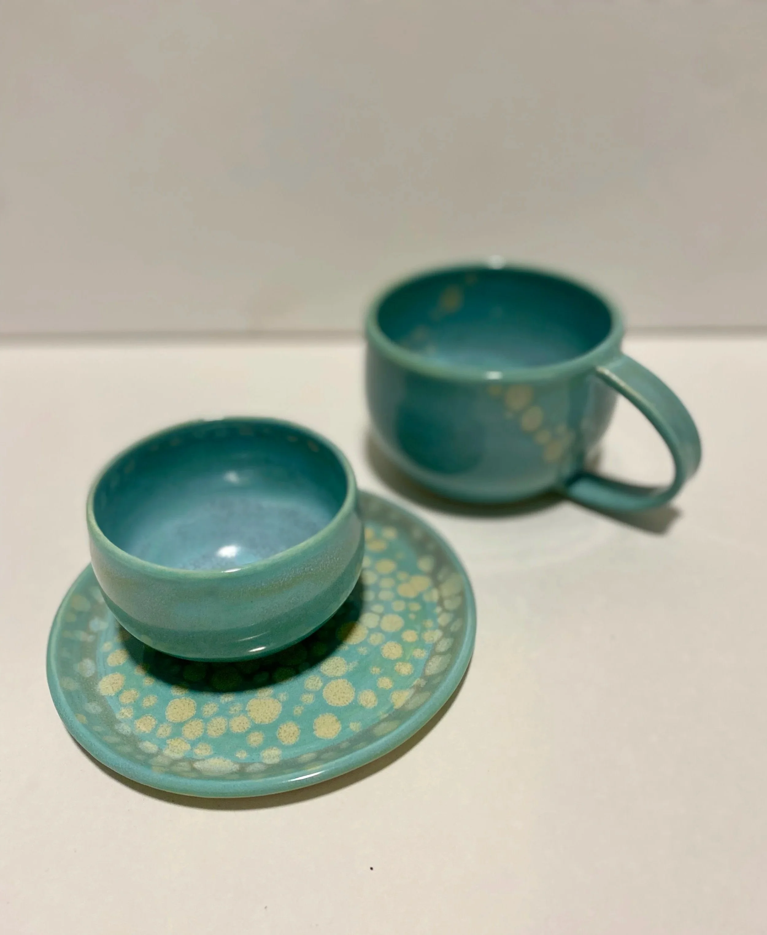 two light blue mugs, one is handleless on a matching plate, all with light circles in the glaze