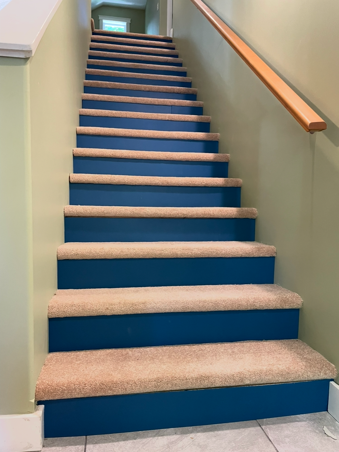 Carpeted staircase with blue risers and beige treads, flanked by light green walls and a wooden handrail on the right.