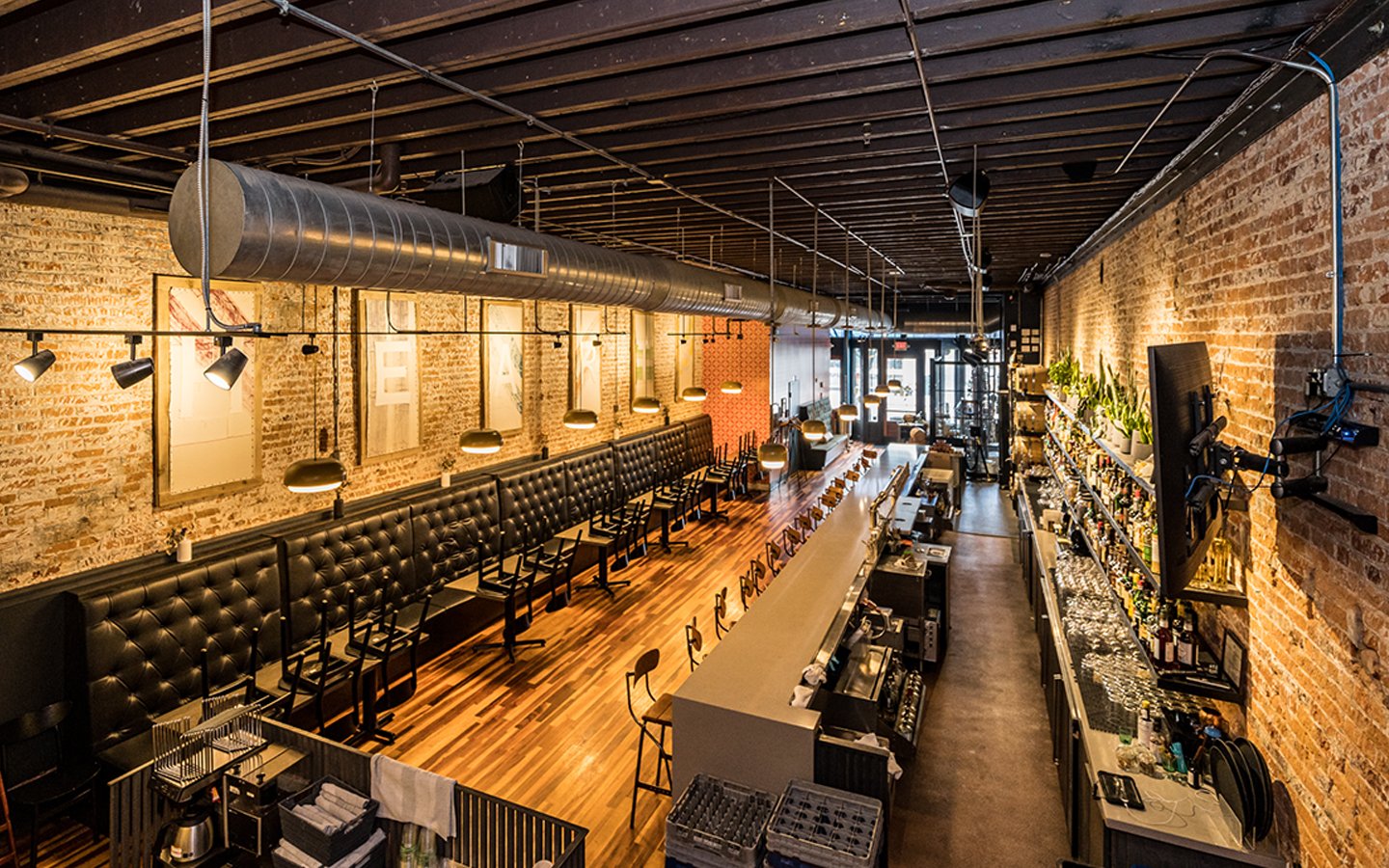 Interior of a modern bar or restaurant with exposed brick walls, wooden floors, black leather banquettes, and a long bar counter on the right side. Hanging pendant lights and framed artwork adorn the walls, with a TV mounted on the brick wall. The space is well-lit and organized with plants and bar equipment.