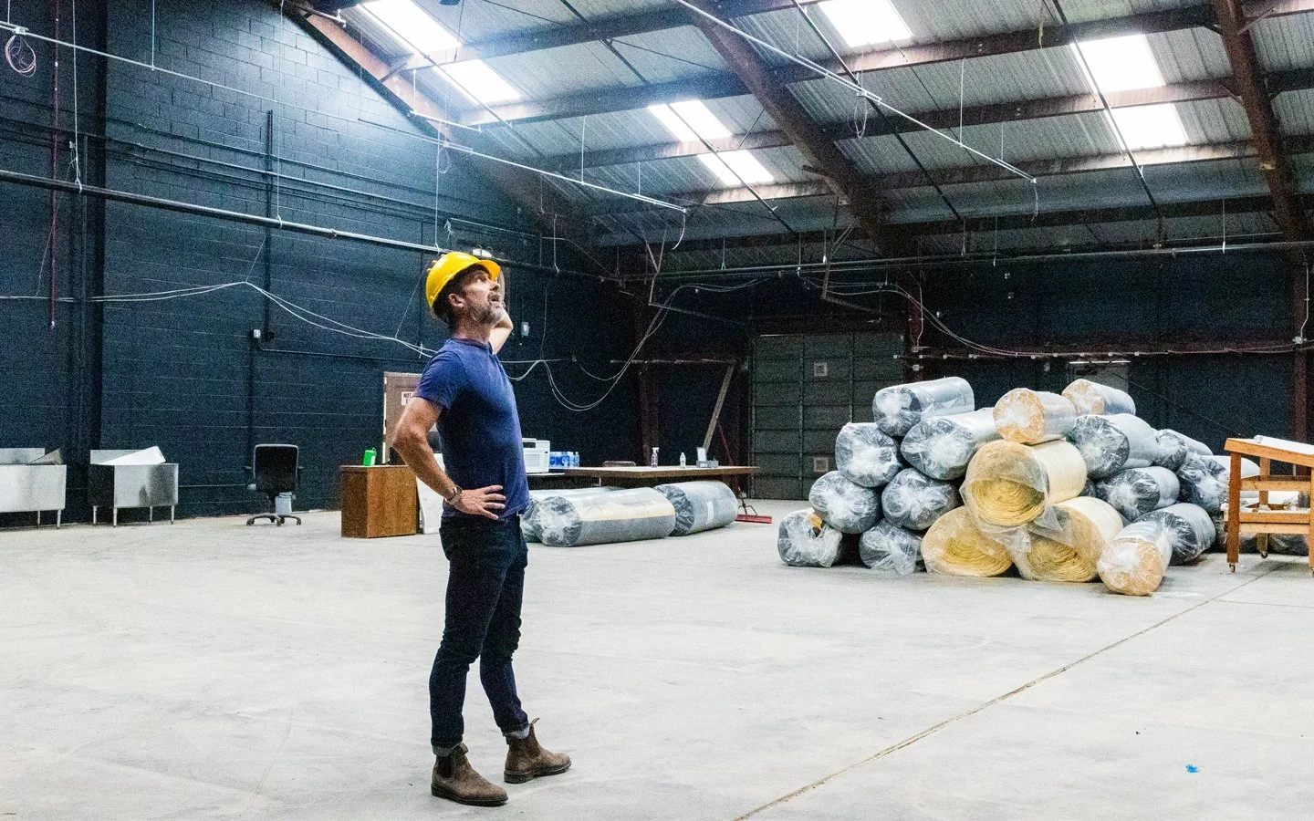 A man wearing a yellow hard hat, blue t-shirt, and brown boots standing with hands on hips inside an industrial warehouse with stacks of insulation rolls and various equipment.