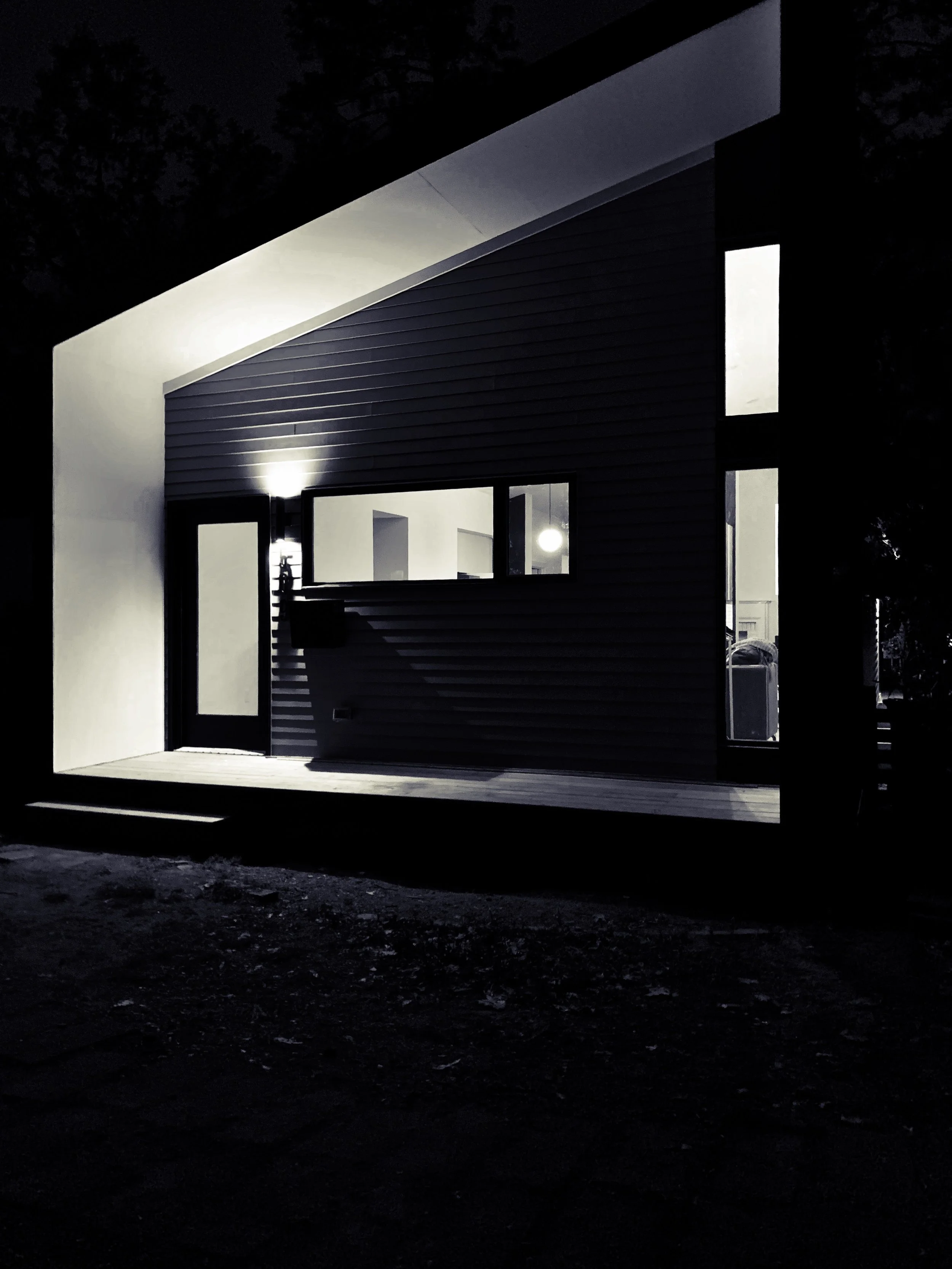 Night view of a modern house with illuminated interior, featuring large windows, a small porch, and exterior lighting, with trees in the background.