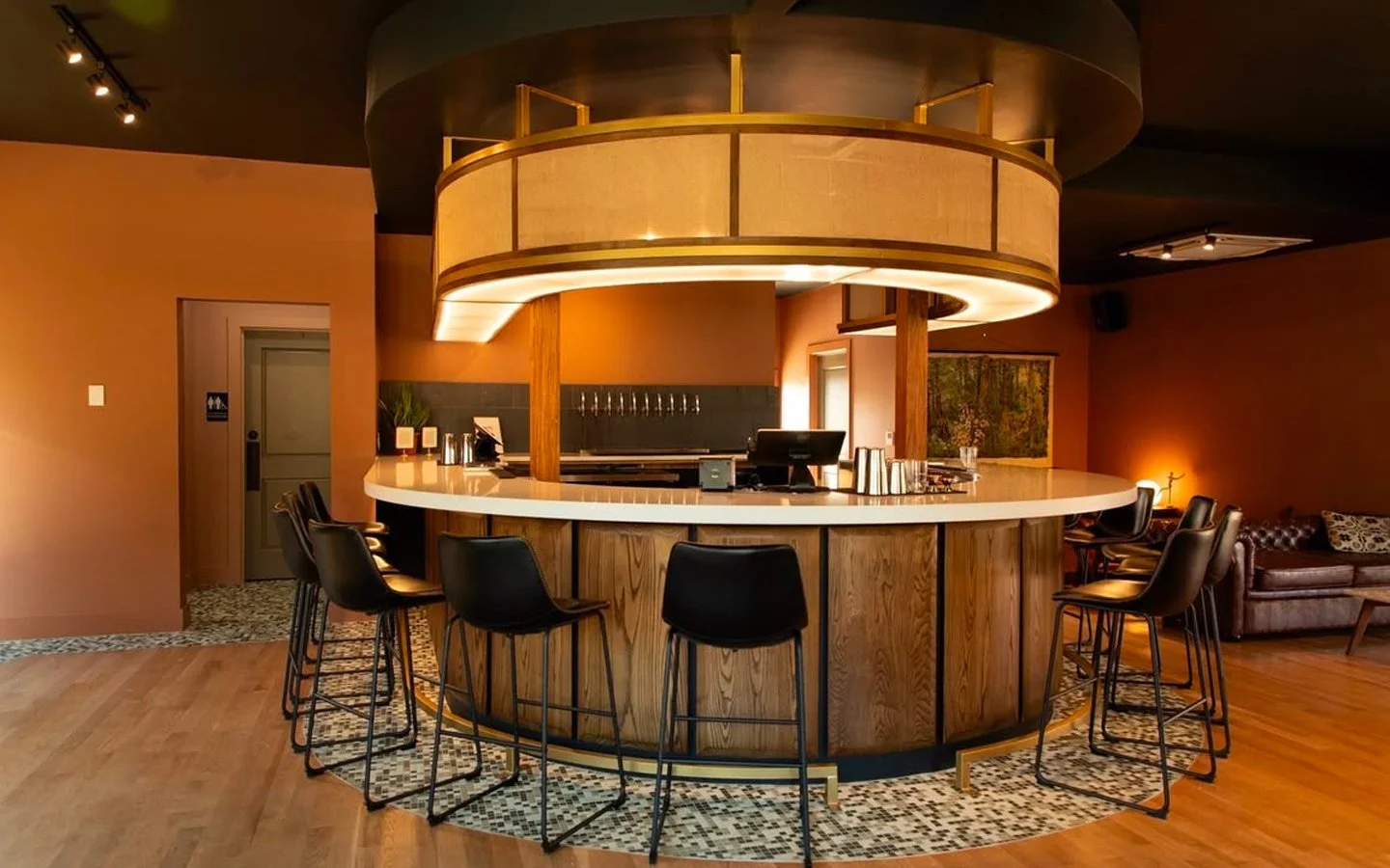 A modern, circular bar with a white countertop and black chairs, illuminated by warm lighting, with a seating area and artwork in the background.