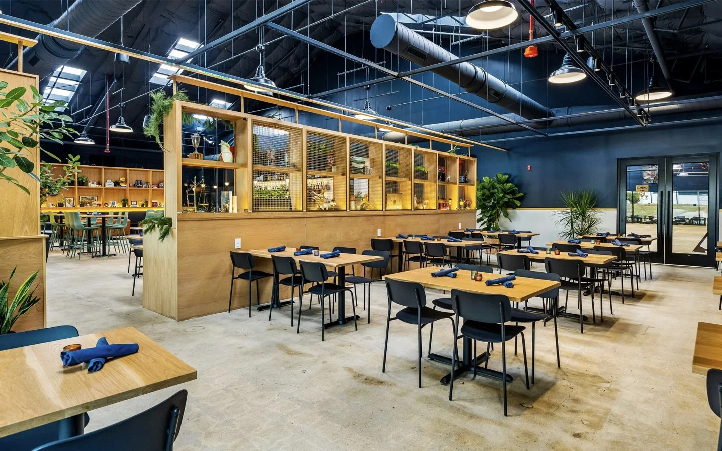Interior of a modern restaurant with wooden tables and black chairs, decorated with plants and set with blue napkins, featuring a high ceiling, black walls, and large windows.