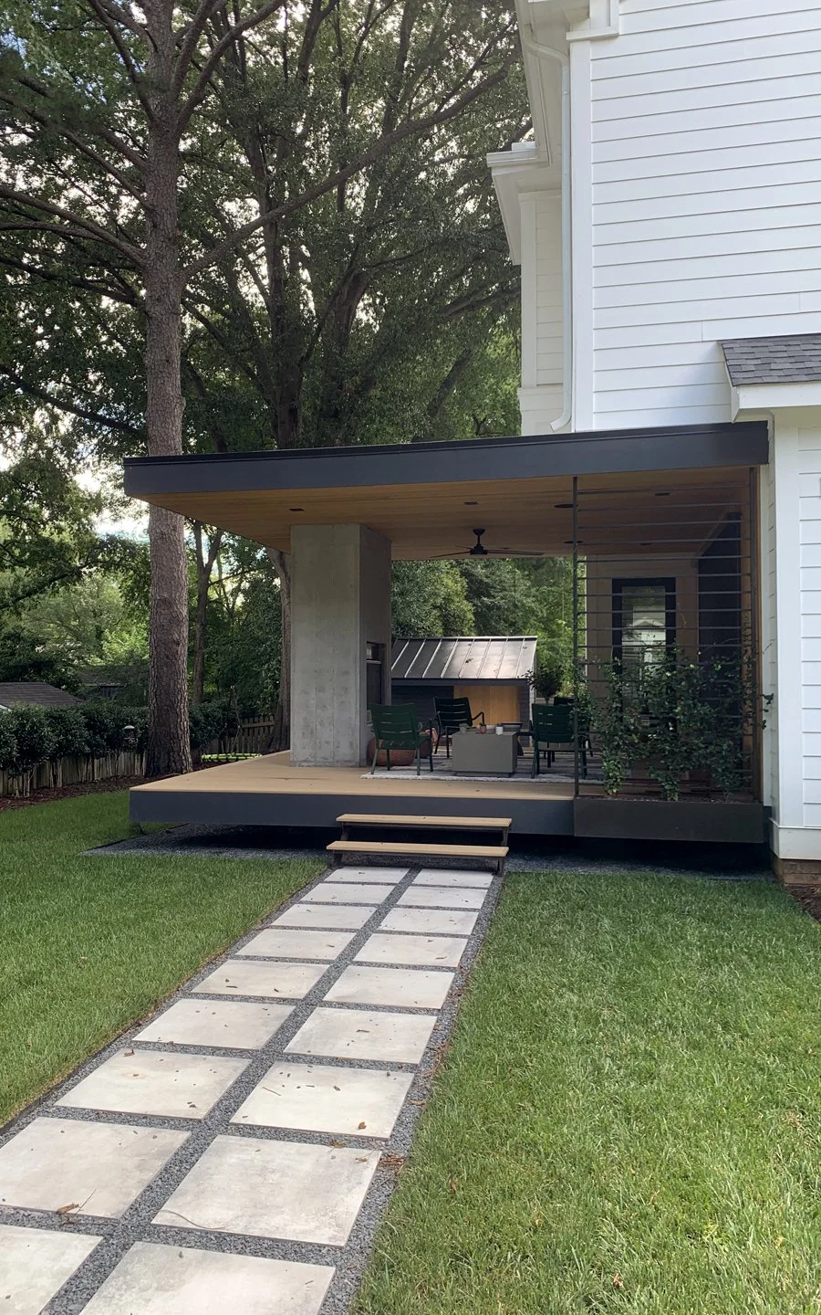 Modern backyard patio with a concrete post, wood ceiling, ceiling fan, outdoor seating area, and a small steps leading up to the patio, surrounded by green lawn and trees.