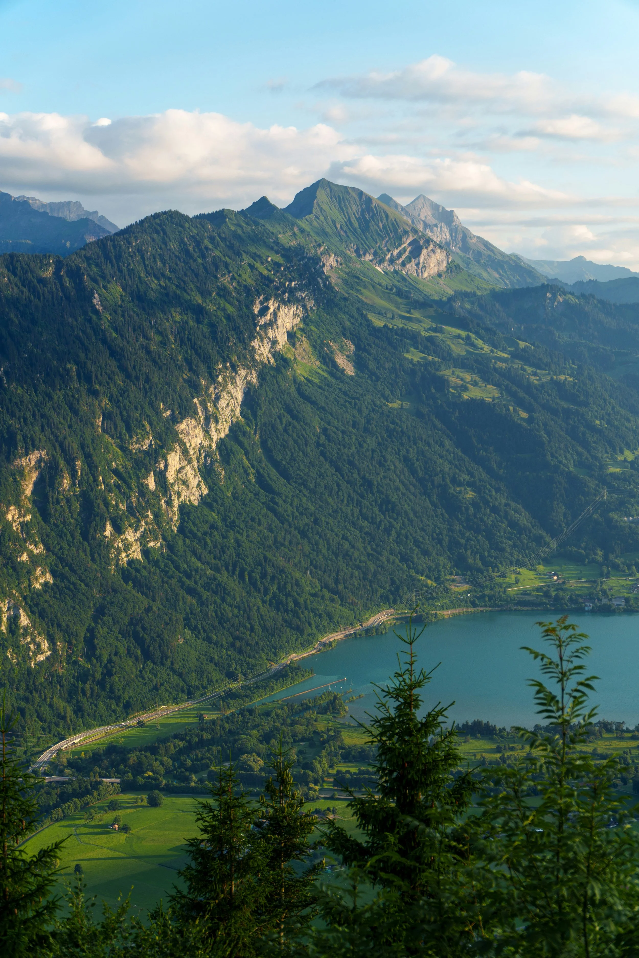 Green mountain range with lush forests and a lake in the foreground, under a partly cloudy sky.