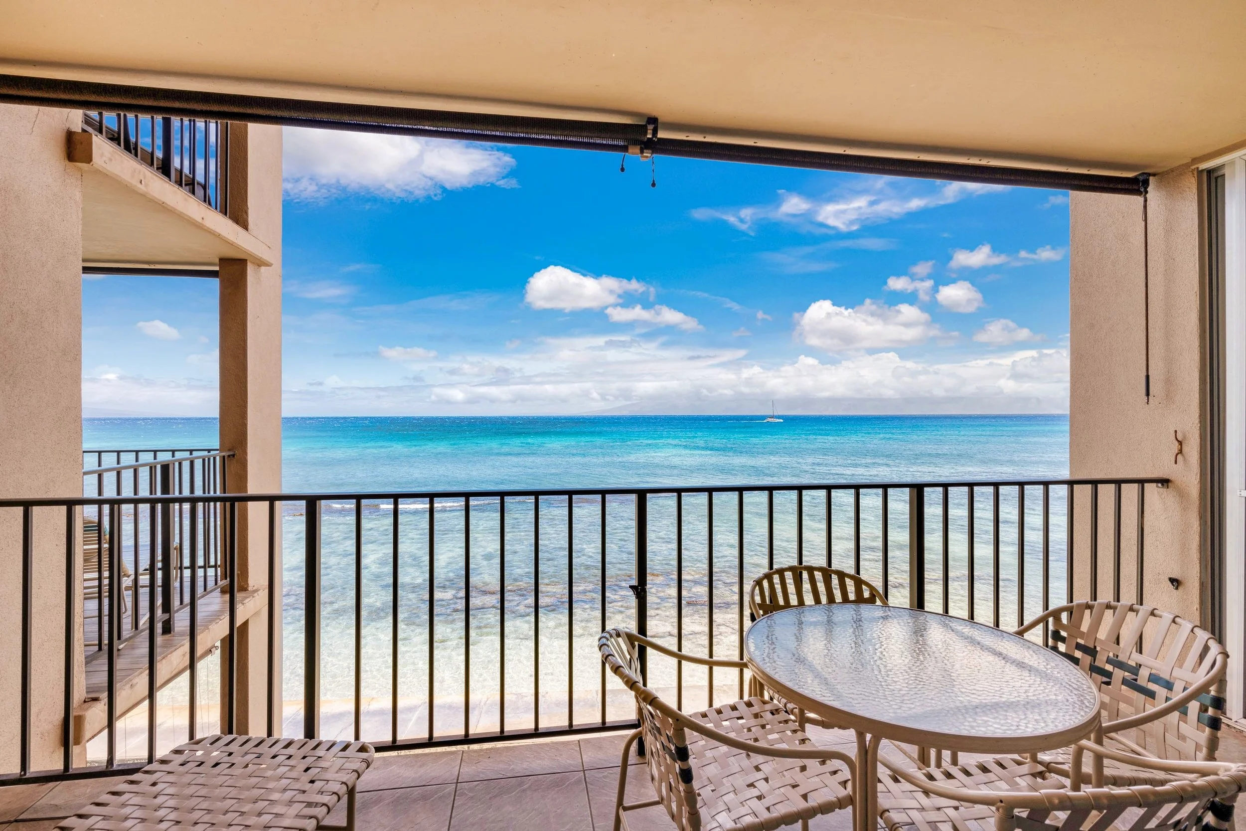 Balcony overlooking the ocean with a glass-top table and four chairs, railing, and building structure in the foreground, blue sky with clouds and sailboat in distance.