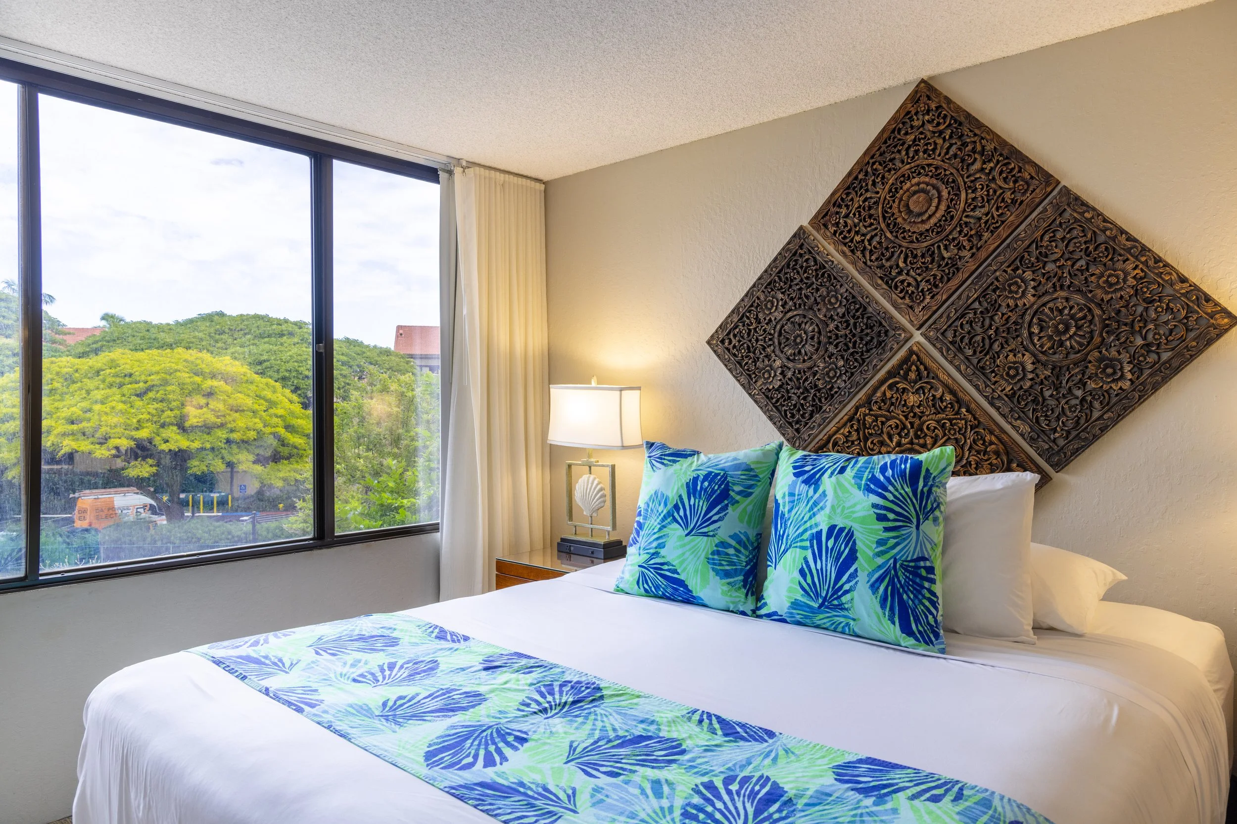 Hotel bedroom with a bed decorated with tropical patterned pillows and runner, a large window showing green trees outside, a bedside lamp, and ornate wooden wall art.