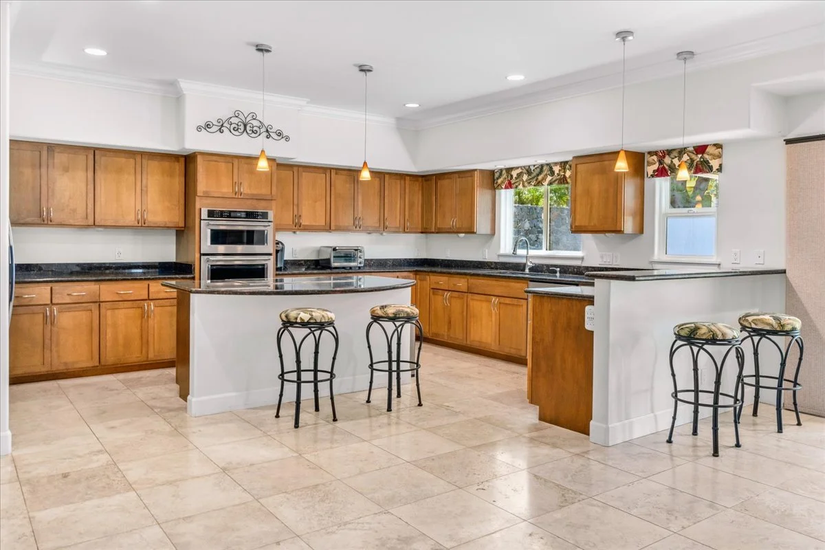 Spacious kitchen with wooden cabinets, black granite countertops, tile flooring, and hanging pendant lights. There are two windows above the sink, and the kitchen features a central island with barstools.