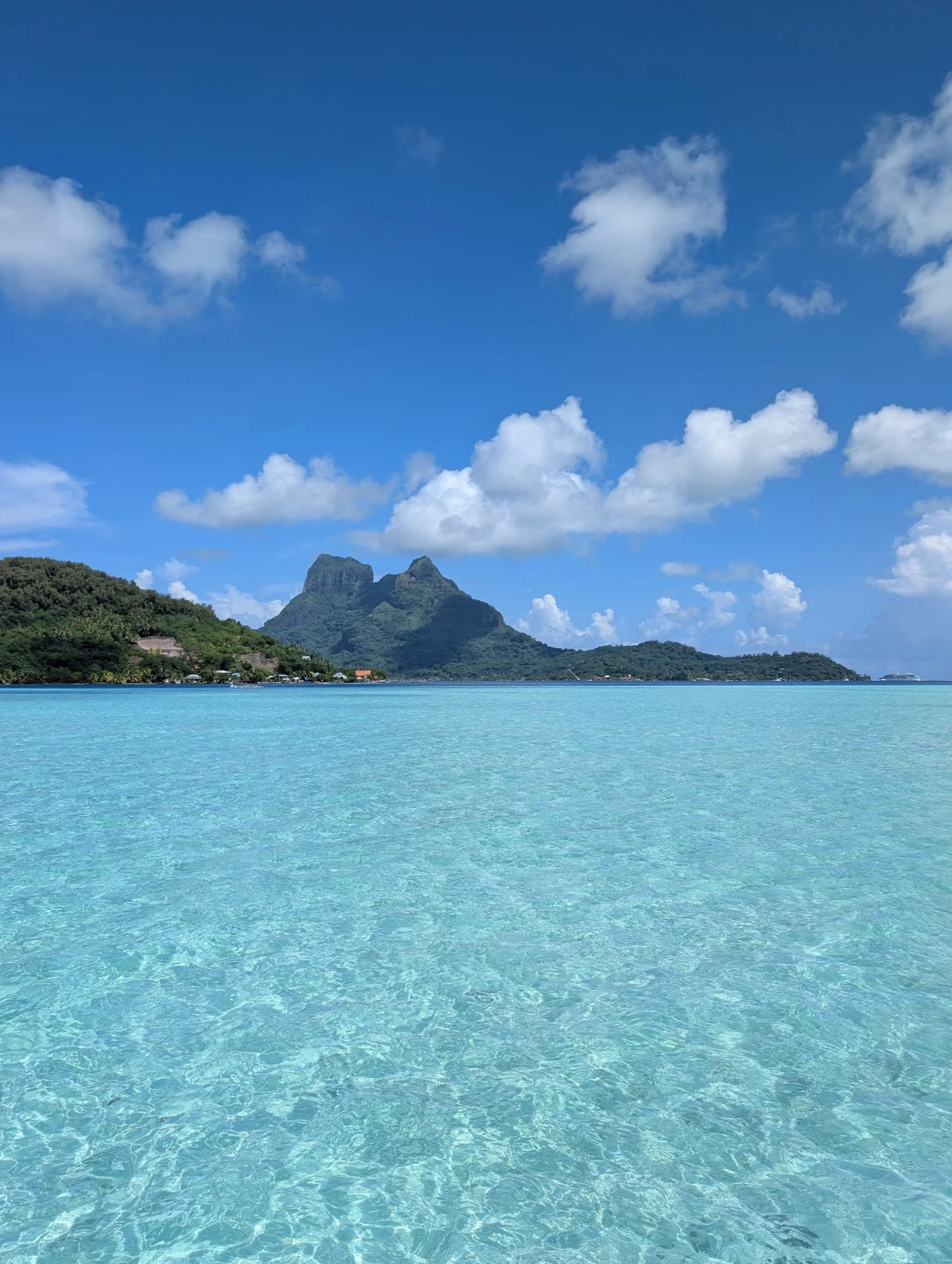 A tropical island with green mountains in the distance, clear turquoise water in the foreground, and a blue sky with white clouds.