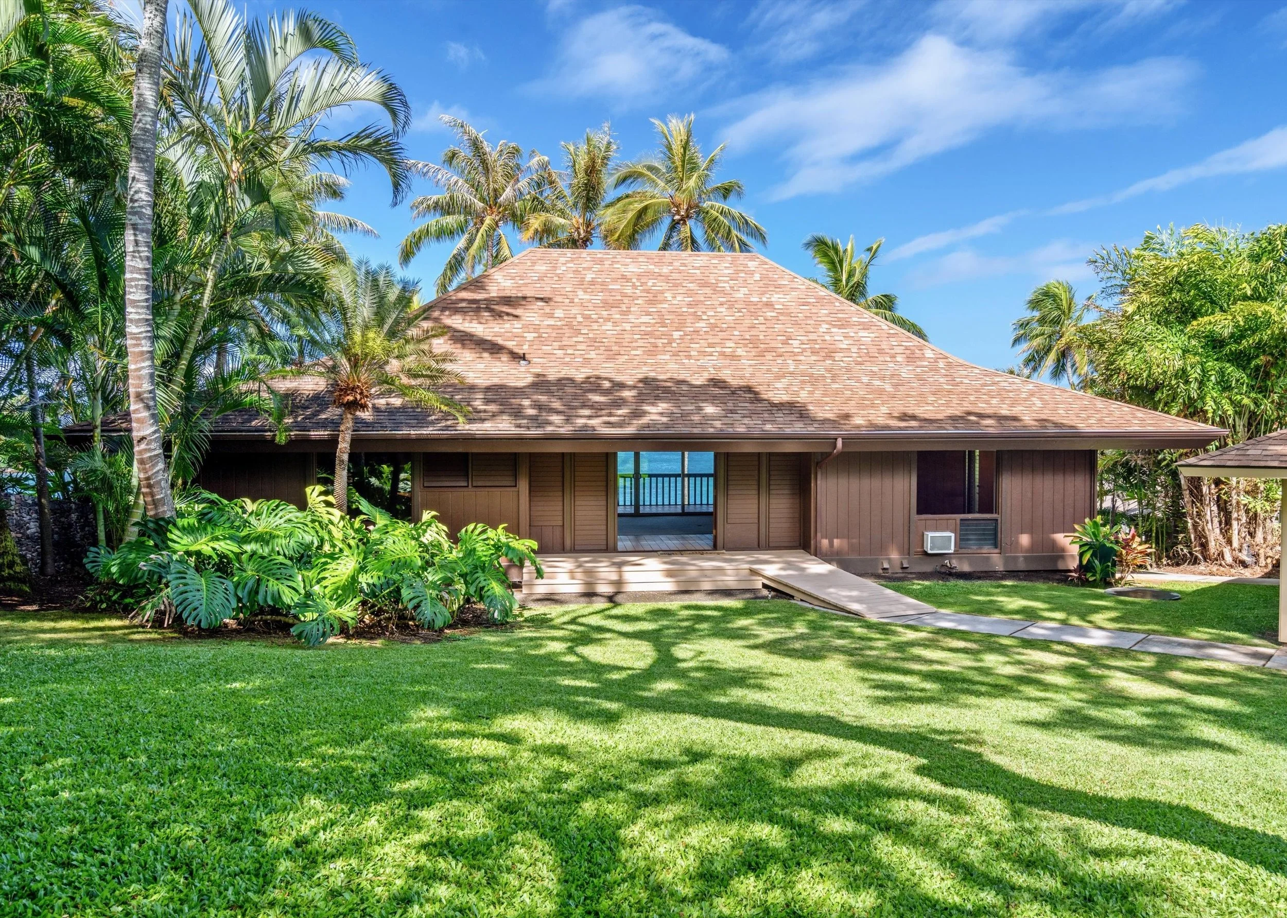 A single-story house in a tropical setting with a brown wooden exterior and a large shingled roof, surrounded by lush green grass, tropical trees, and plants, with a view of the ocean through an opening in the house