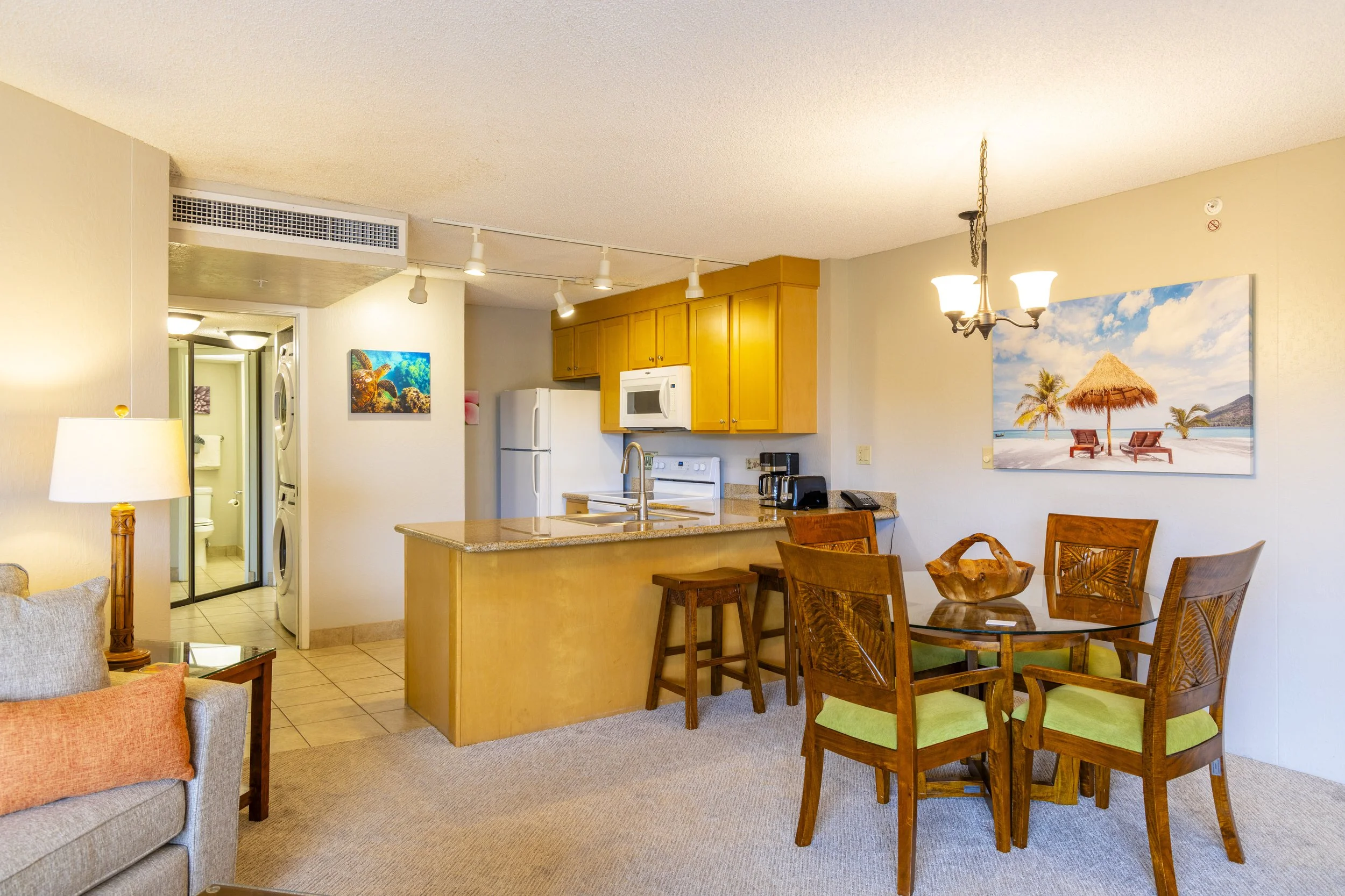 Living room and kitchen area in a vacation rental with tropical decor, includes a dining table with four chairs, a kitchen island, and a view of a laundry area and appliances.