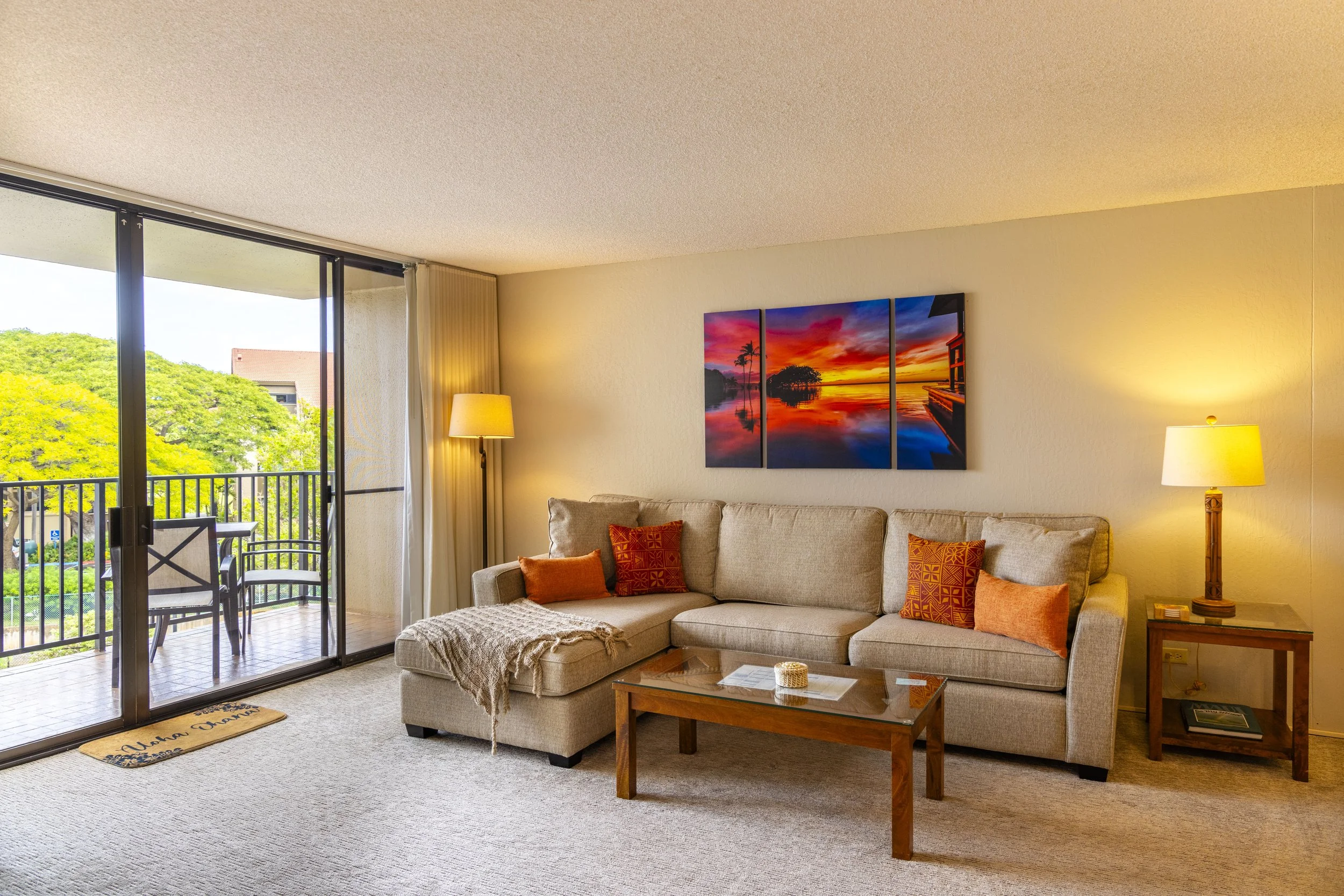 Living room with beige sectional sofa, colorful throw pillows, wooden coffee table, and sliding glass door leading to a balcony. There are two table lamps with warm yellow light, a framed sunset landscape painting on the wall, and greenery visible outside.