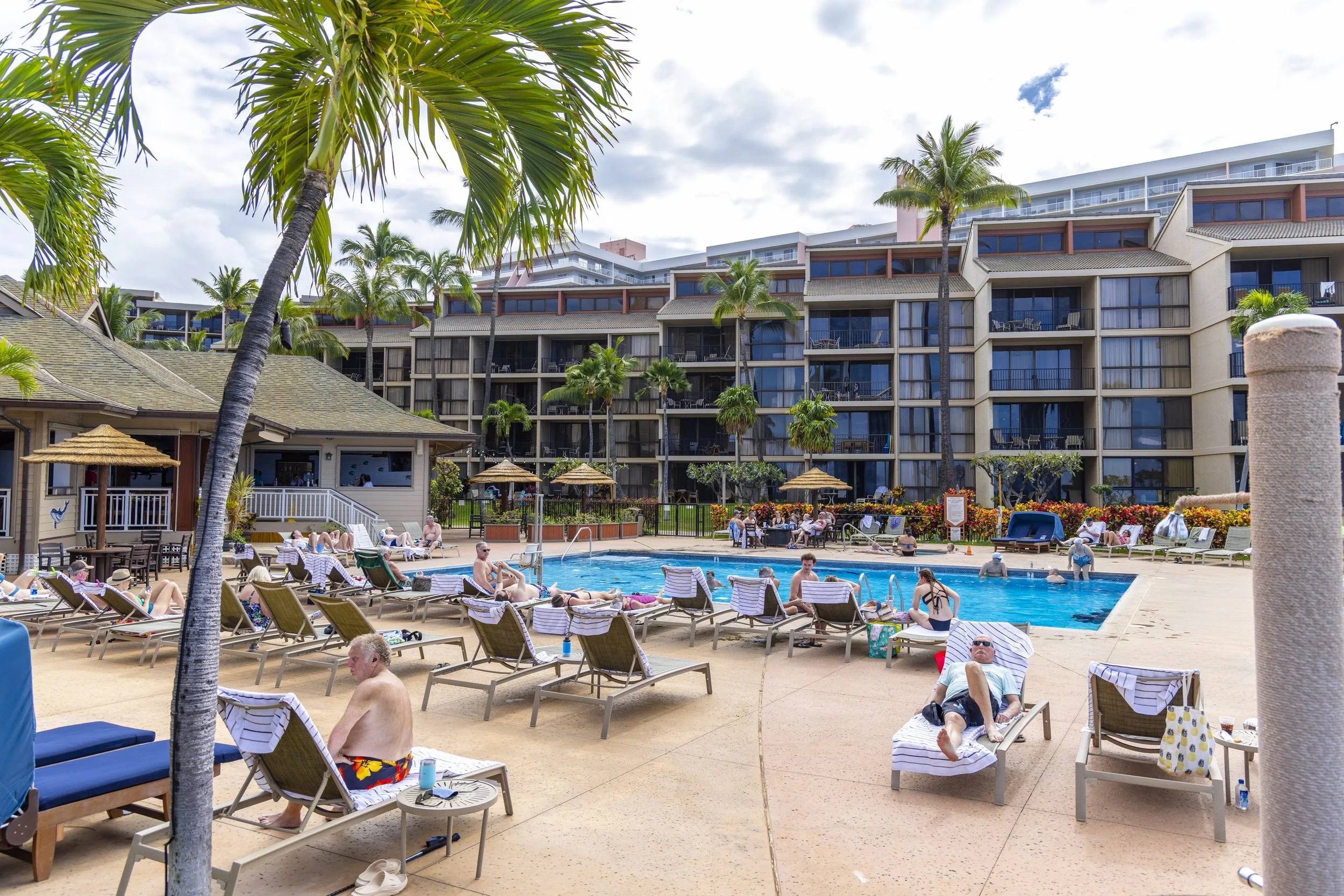 People relaxing on lounge chairs and in the pool area at a resort with palm trees and a large multi-story building in the background.