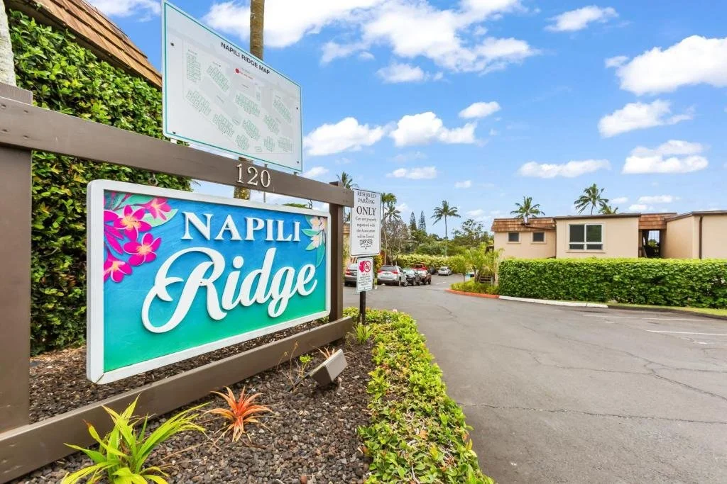 Sign indicating the entrance to Napili Ridge with a parking lot and residential buildings in the background under a partly cloudy sky.