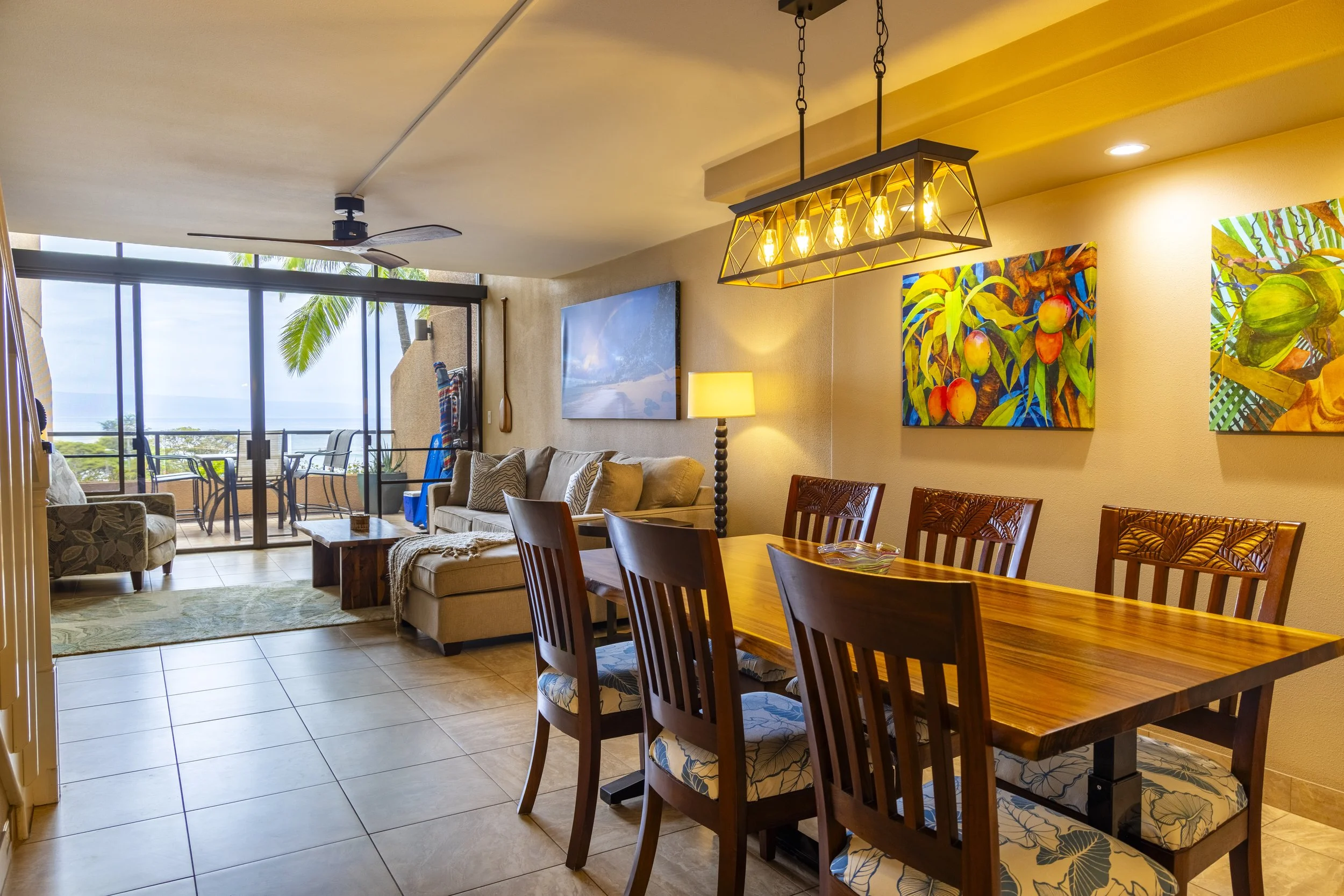 Living room and dining area with sliding glass door leading to balcony with outdoor seating, tropical ocean view, and colorful tropical themed artwork on the wall.