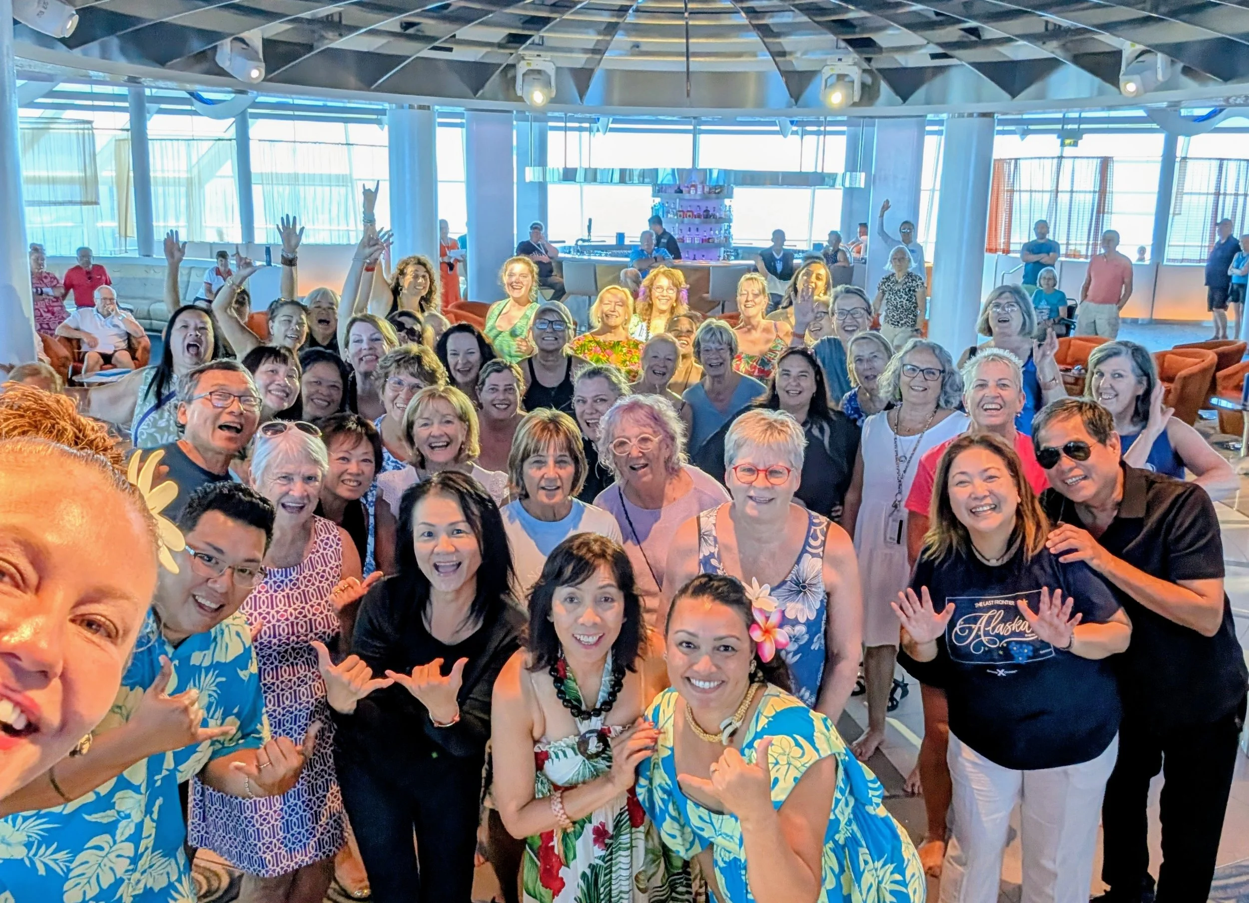 Group of smiling adults at a social gathering on a cruise ship, with tropical-themed clothing and flowers, in a lounge area with large windows and modern decor.
