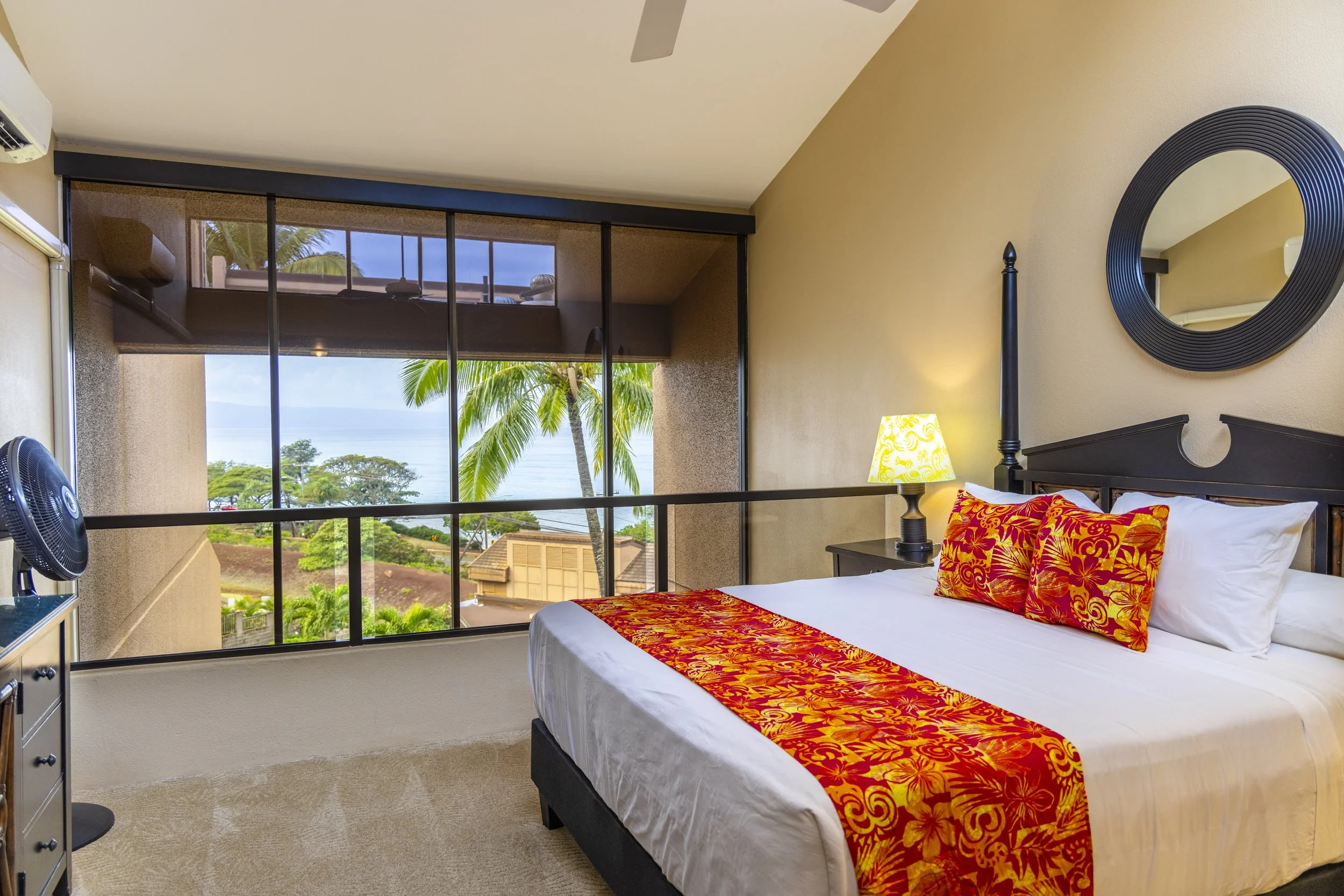 Hotel room with a large window showing palm trees and ocean view, a bed with white sheets, red and yellow patterned throw pillow and bed runner, a black headboard, a bedside lamp, a mirror, an air conditioning unit, and a small black fan.