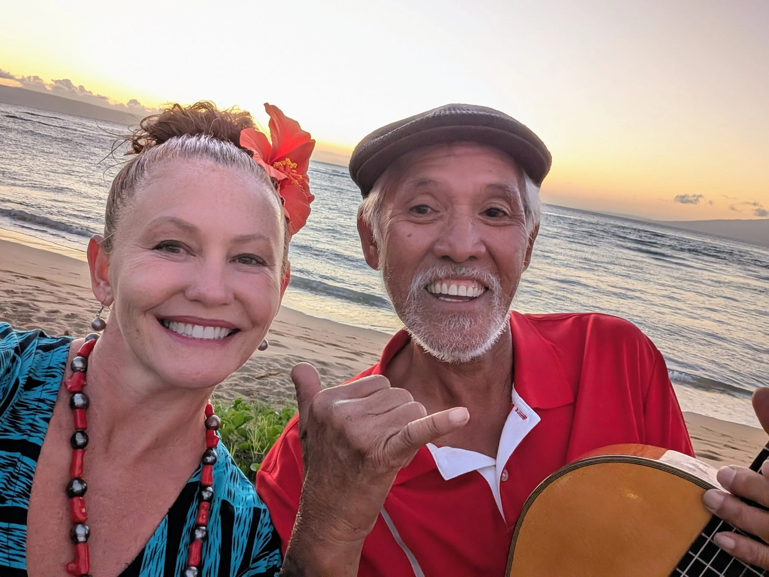 Smiling woman with red flower hair accessory and beaded necklace taking a selfie with smiling man wearing a cap, playing guitar on a beach at sunset.