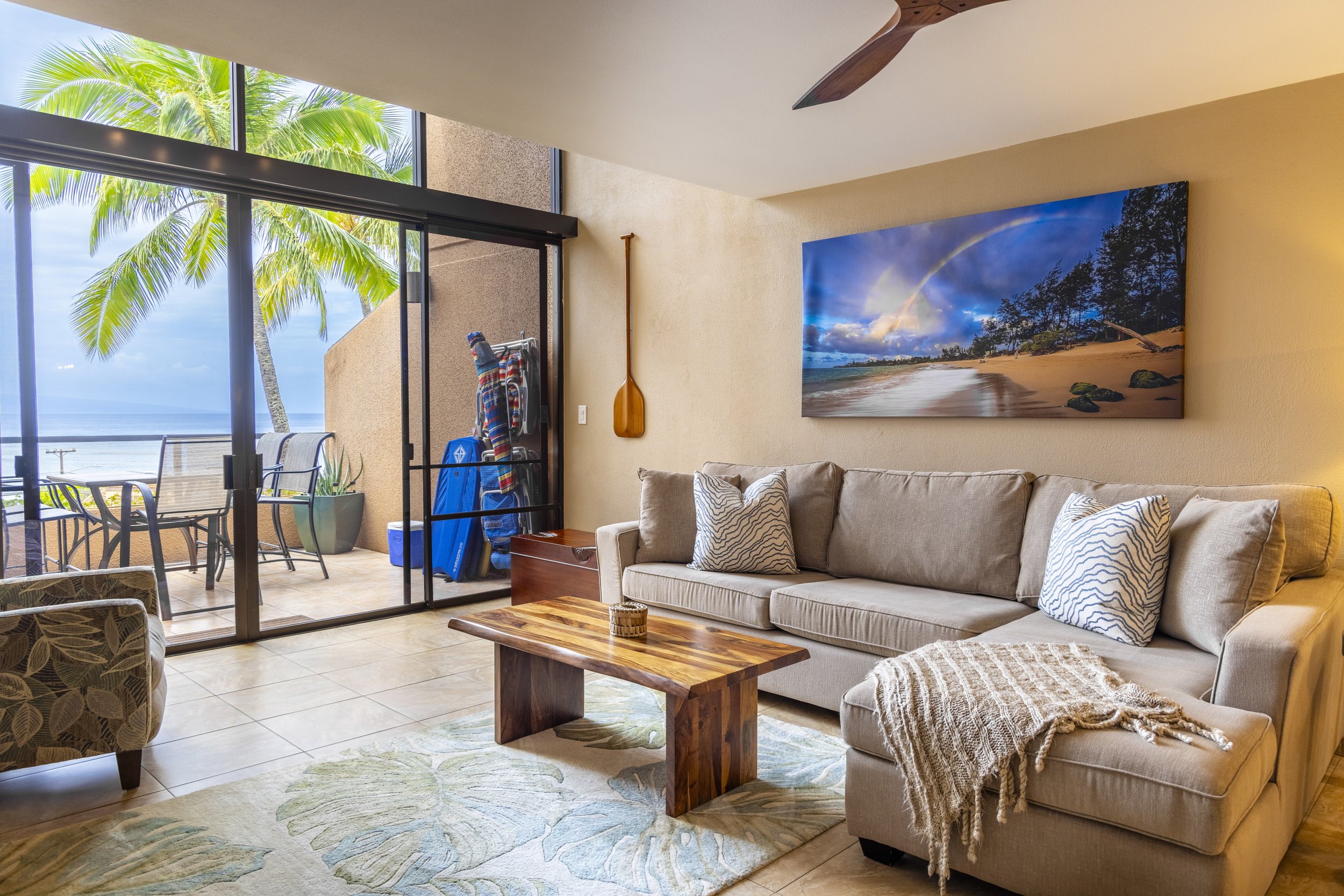 Living room with beige couch, decorative pillows, wooden coffee table, large wall art of a beach scene with blue sky and rainbow, sliding glass door opening to balcony with outdoor seating and view of palm trees and ocean
