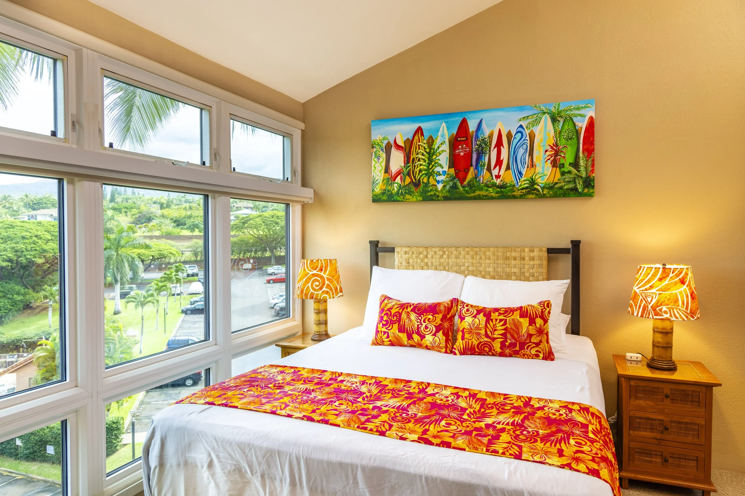 Hotel room with tropical decor, bed with colorful floral and leaf-patterned bedspread and pillows, large windows showing palm trees and parking lot outside, paintings of surfboards and tropical plants on wall, two wooden nightstands with lamps.