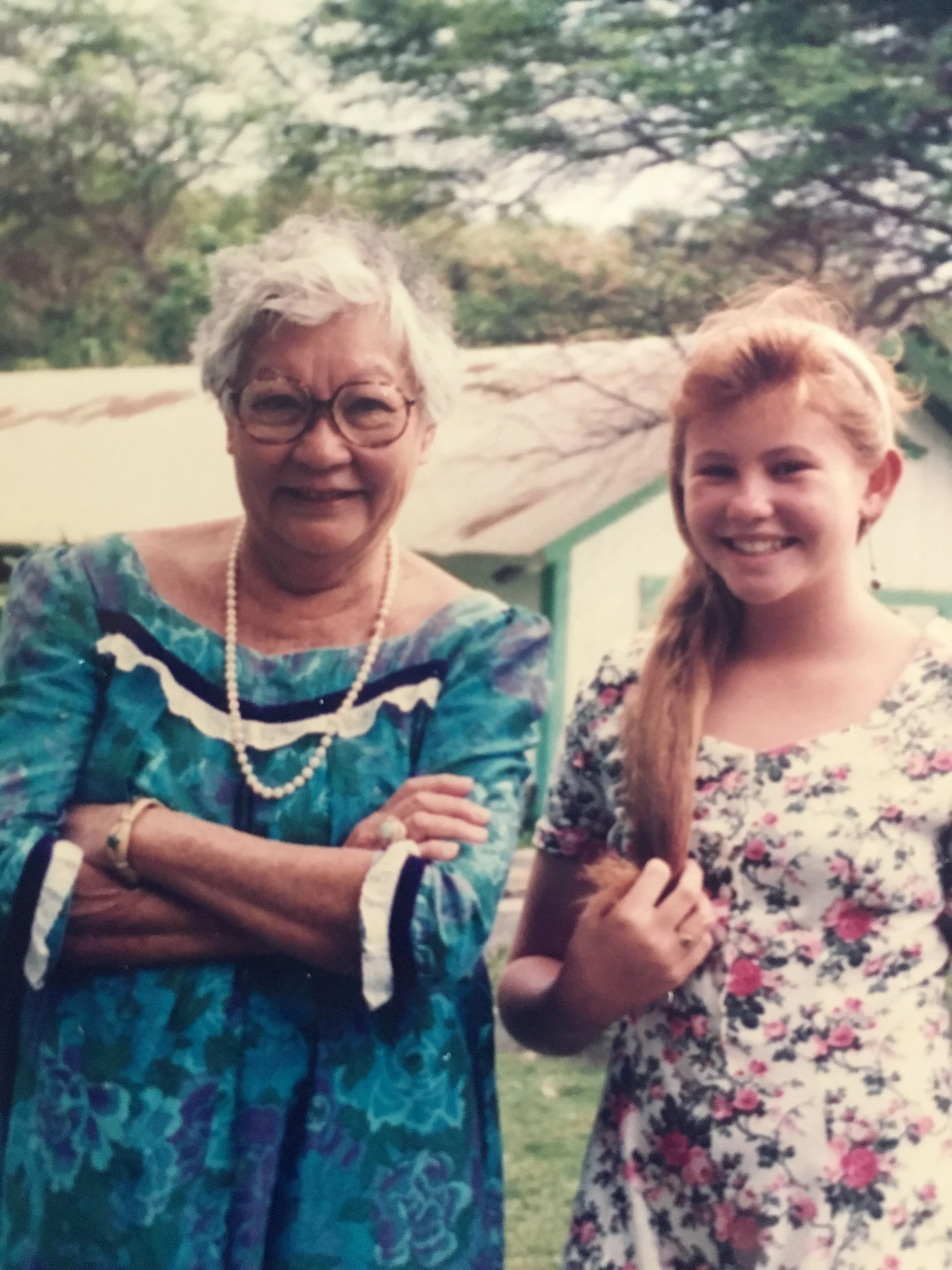 A photo of an elderly woman with white hair and glasses standing next to a young girl with long red hair outdoors. Both are smiling and wearing floral dresses.