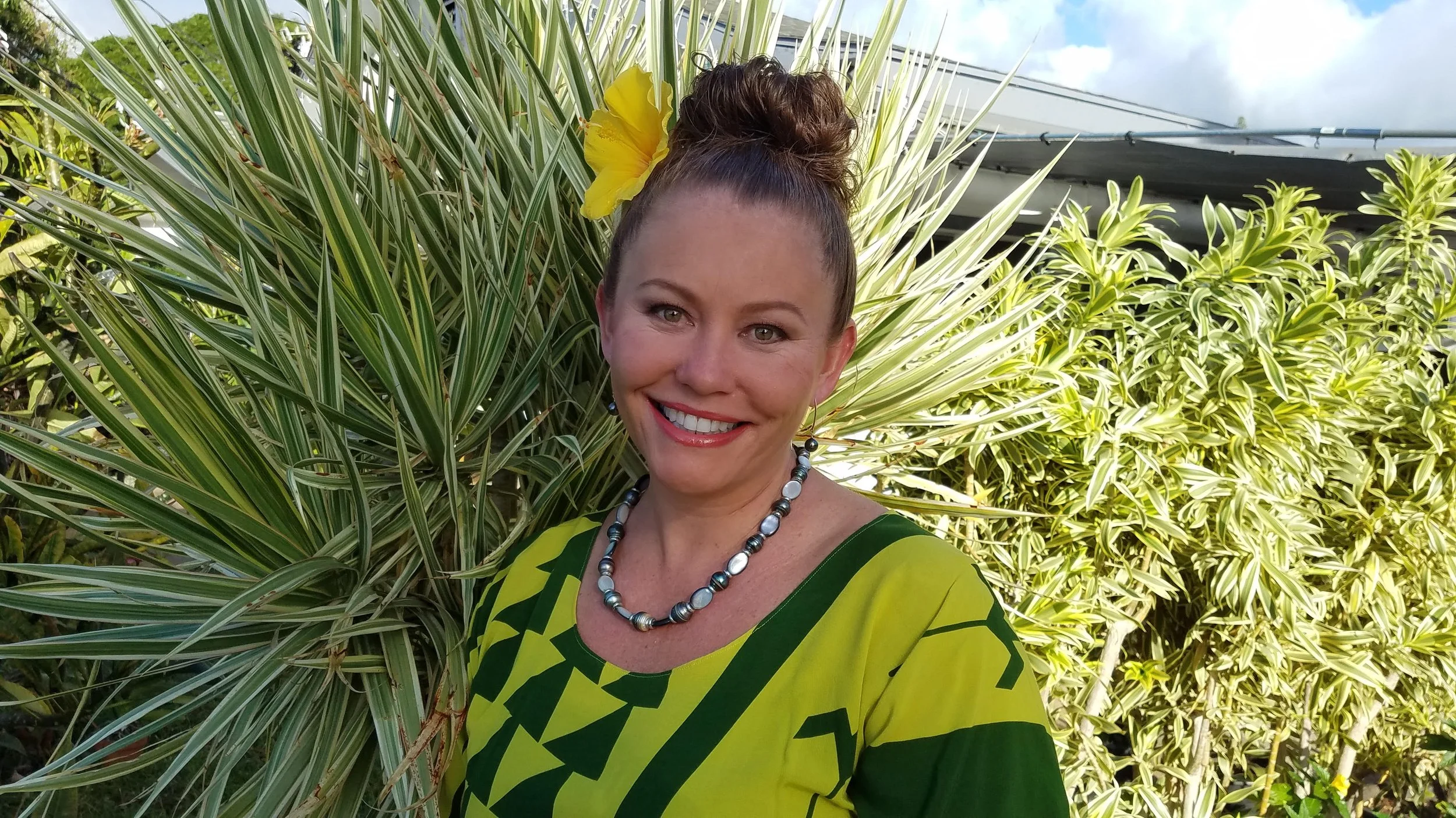 A smiling woman with a tan complexion, wearing a green and yellow patterned top, a multicolored beaded necklace, and earrings, standing outdoors among green tropical plants, with a yellow flower in her hair.
