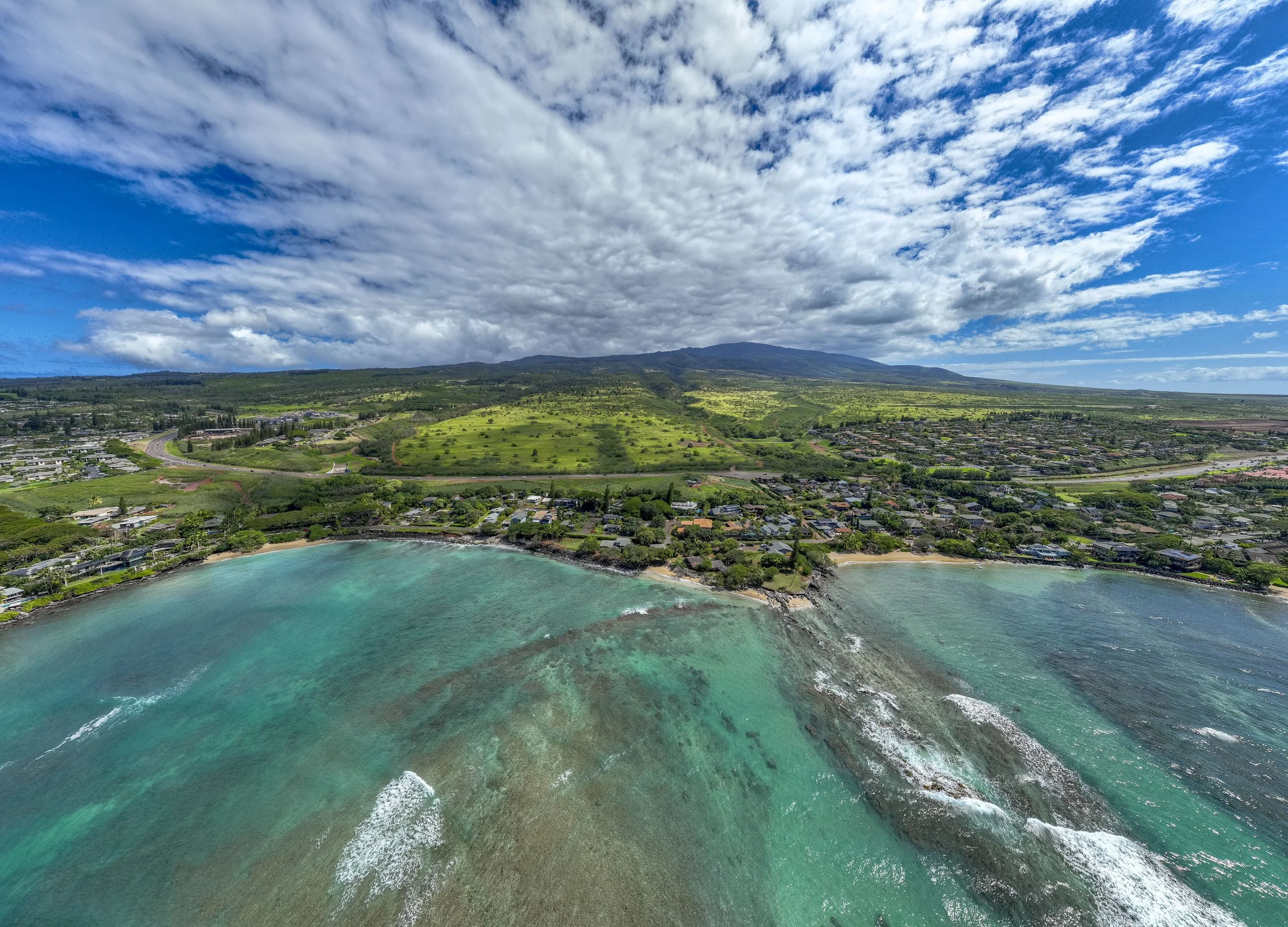 Aerial view of a coastline with turquoise water, residential houses, green hills, and a mountain under a partly cloudy sky.
