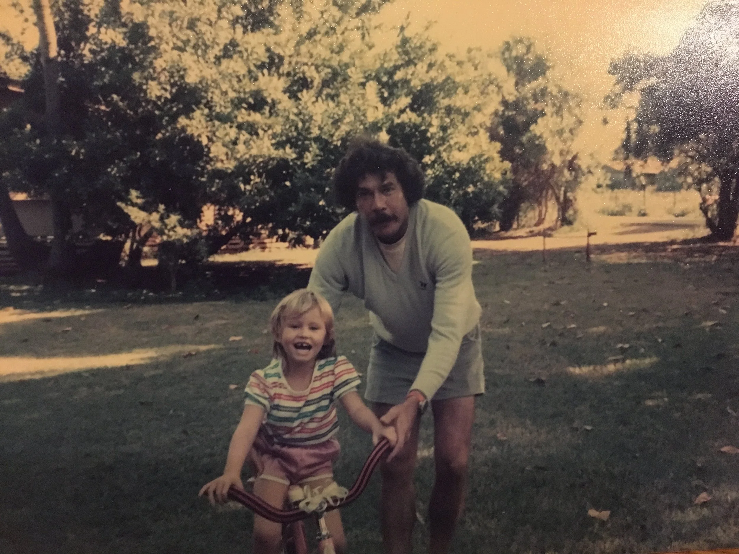 A man with curly dark hair and a thick mustache, wearing a light-colored long-sleeve shirt and shorts, is helping a young girl with blonde hair, wearing a striped shirt and shorts, ride a bicycle outdoors on a grassy area with trees in the background during daytime.