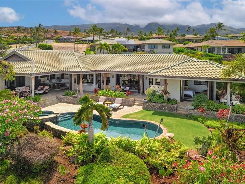 A backyard view of a house with a tiled roof, a swimming pool, and lush tropical landscaping, including palm trees and colorful flowering plants, with neighboring houses in the background and mountains under partly cloudy skies.