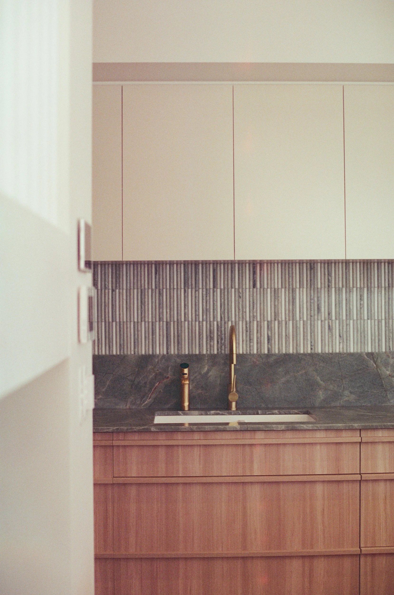 Minimalist kitchen with a dark marble countertop, brass faucet, wood cabinets, and beige wall cabinets.