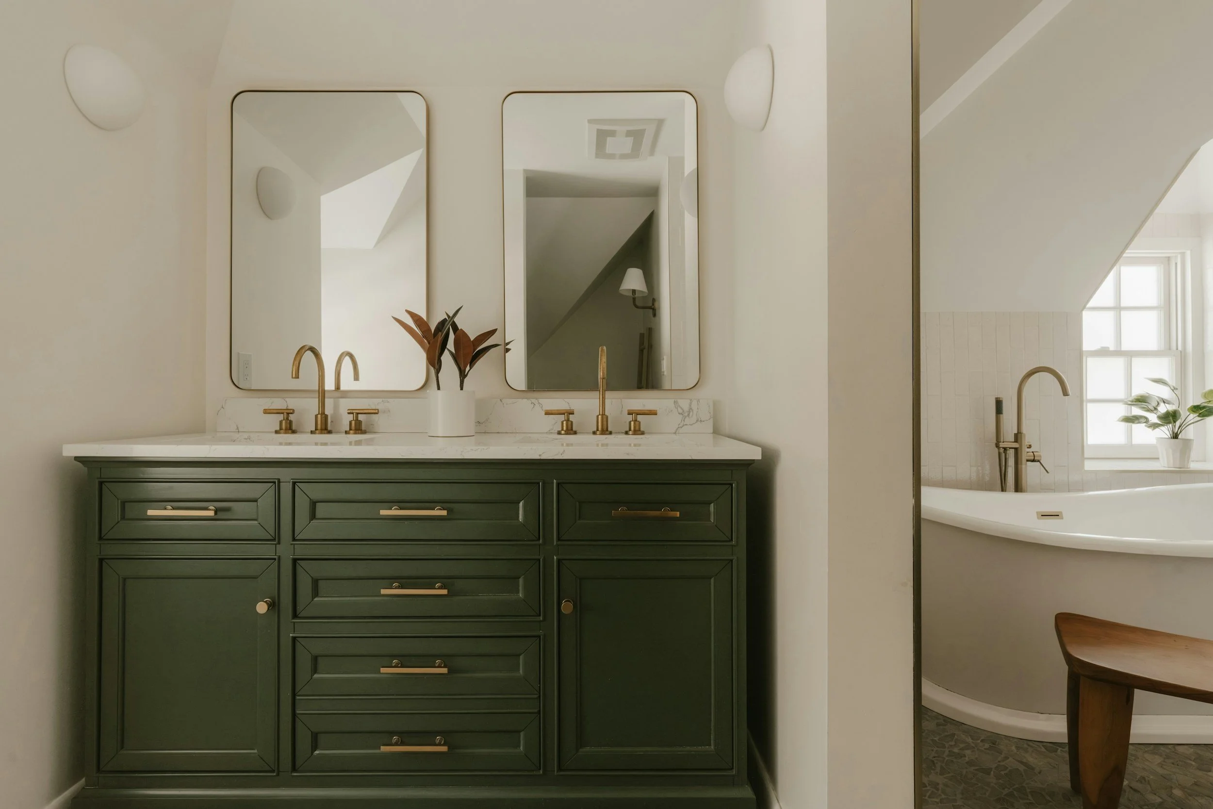 Bathroom vanity with green cabinet, white marble countertop, two mirrors, gold fixtures, and a vase with plants. In the background, a bathtub with a gold faucet, a window, and a wooden stool are visible.
