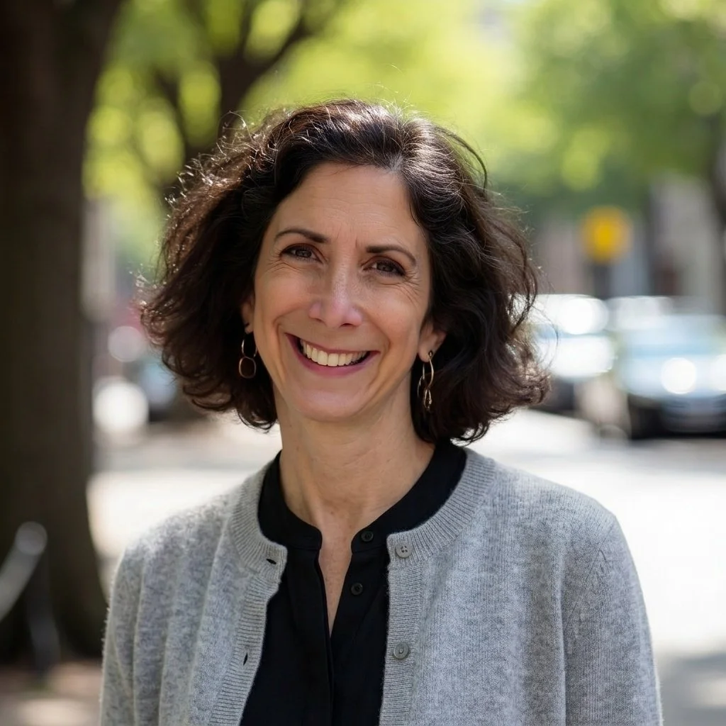 A smiling woman with curly brown hair standing outdoors on a sunny day with blurred trees and parked cars in the background.