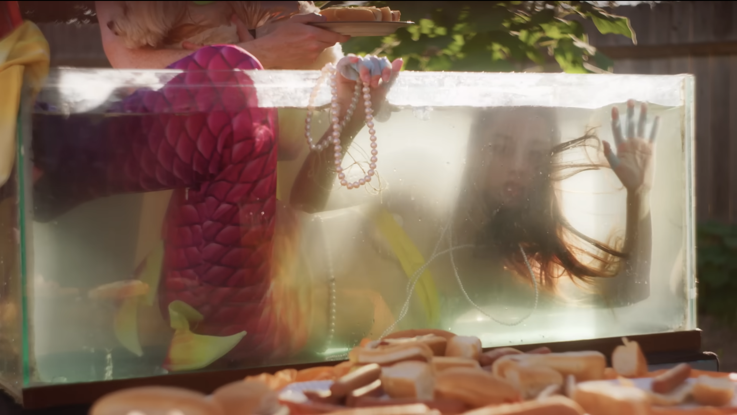 A woman is submerged in a glass tank filled with water, holding a string of pearls in her hand and waving with her other hand, while sunlight illuminates her hair and the water surface. A person outside the tank is holding a tray of food in the background.