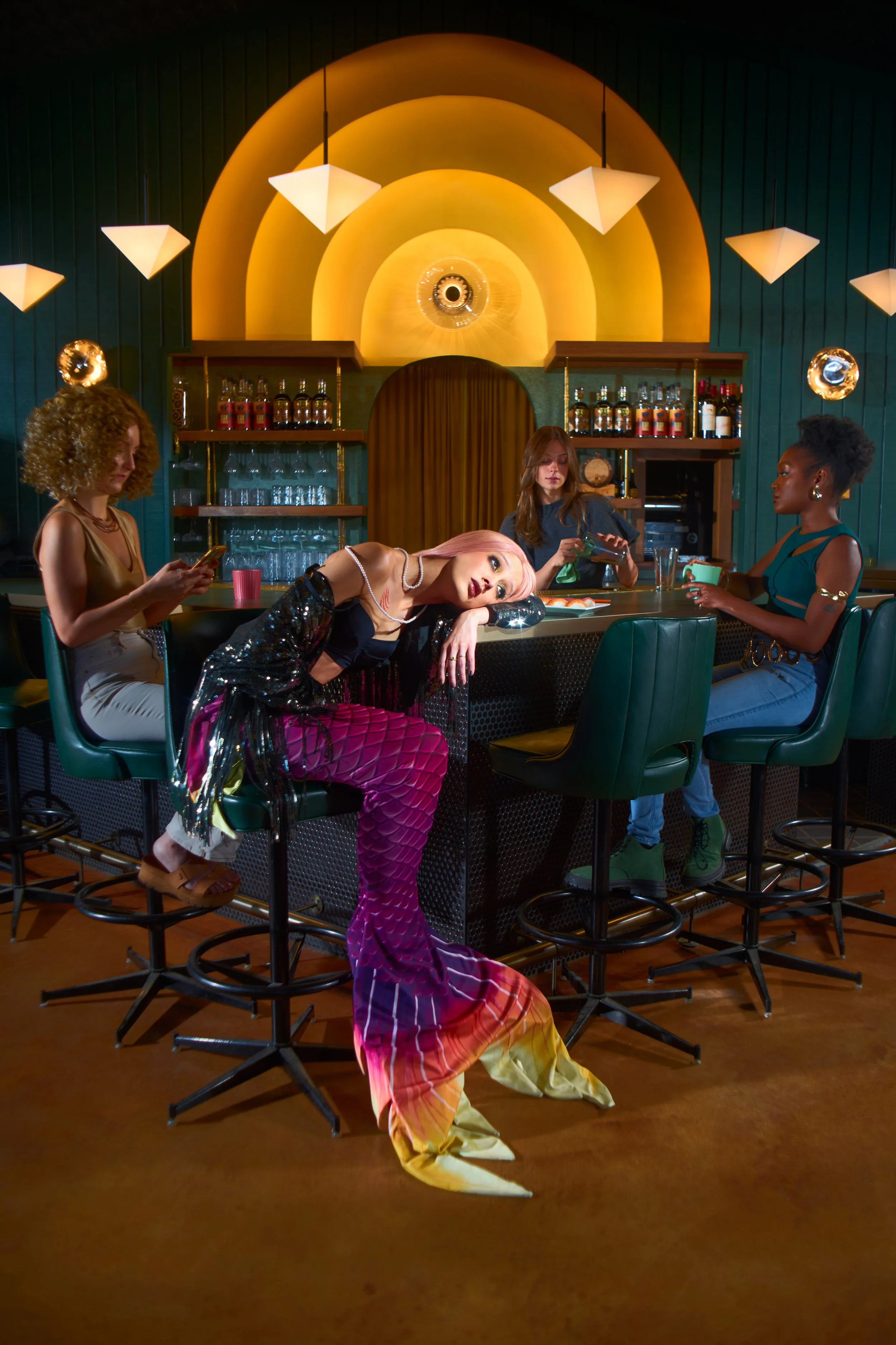 A group of four women sitting at a bar with colorful lighting and decor, one woman in the center leaning on the counter with her head resting on her arm, dressed in vibrant, colorful mermaid-themed attire.