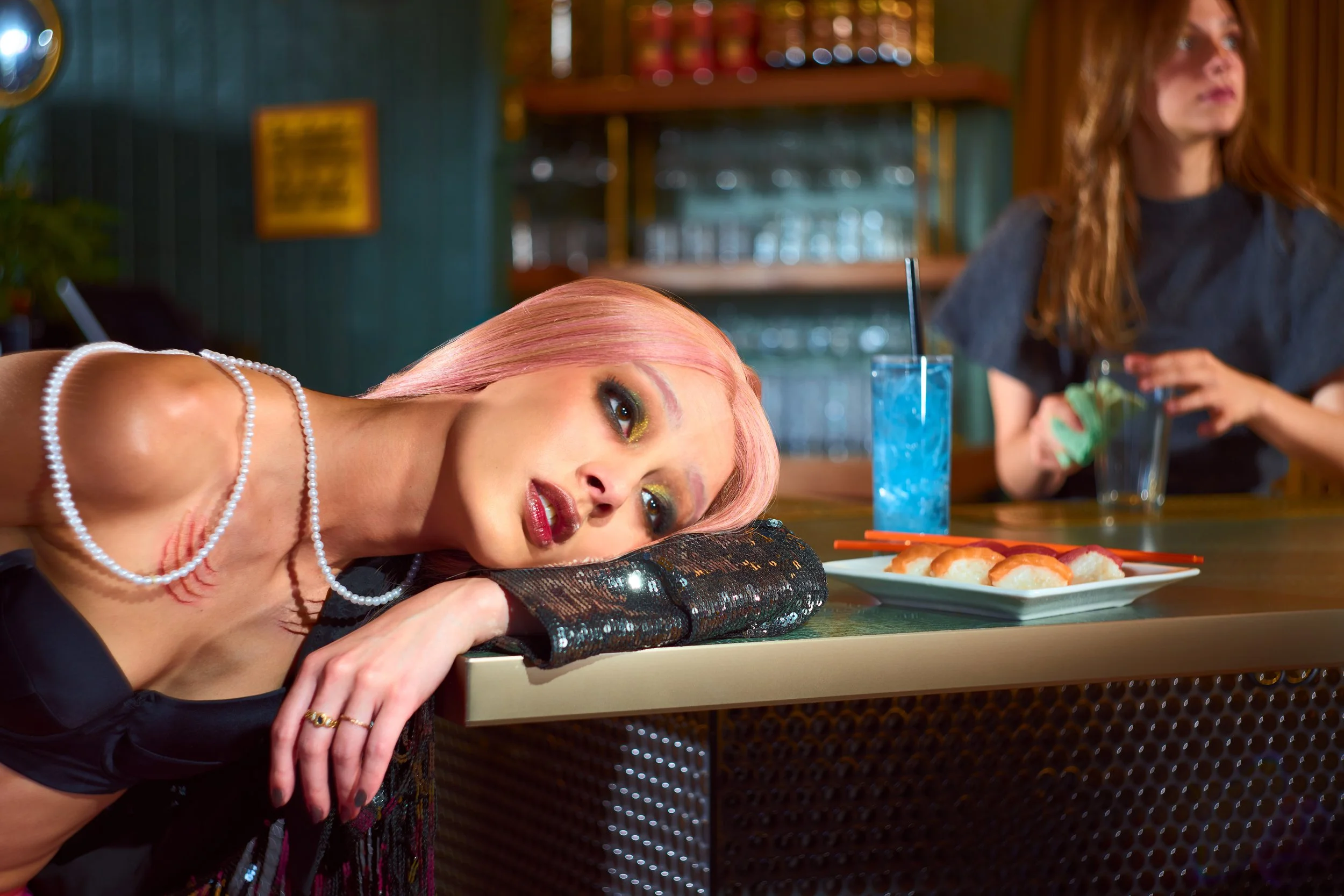 A woman with pink hair wearing makeup and pearls resting her head on her arms at a bar counter with a blue cocktail, sushi, and another person in the background
