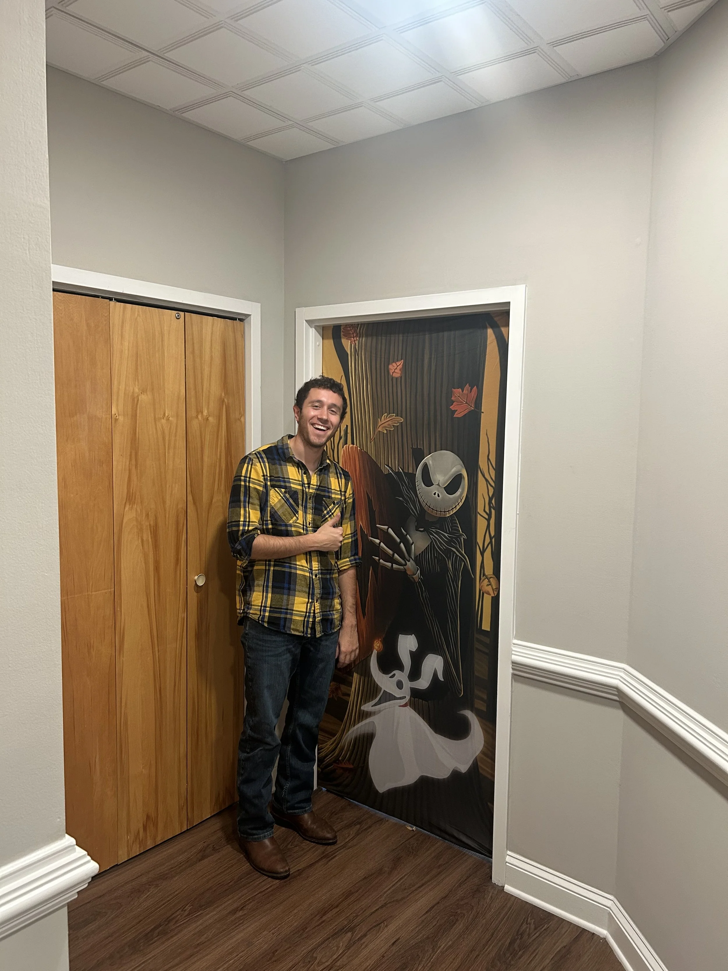 A smiling man wearing a yellow plaid shirt and jeans stands indoors next to a Halloween-themed door with a skeleton illustration. The room has a closet with wooden doors and gray walls.