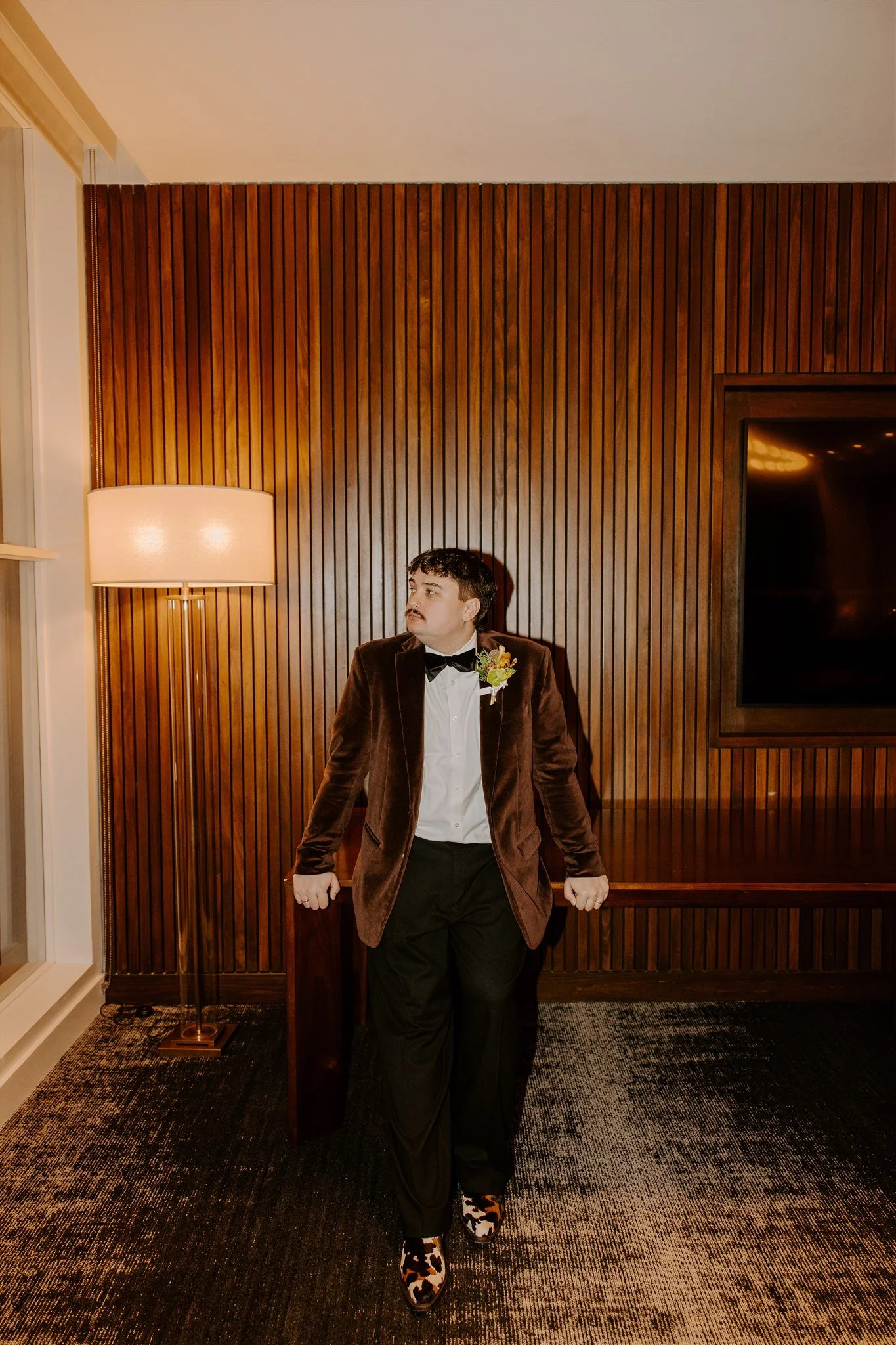 A man in a tuxedo with a brown velvet blazer, black bow tie, and floral-patterned shoes standing in a room with wooden panel walls and a large flat-screen TV.
