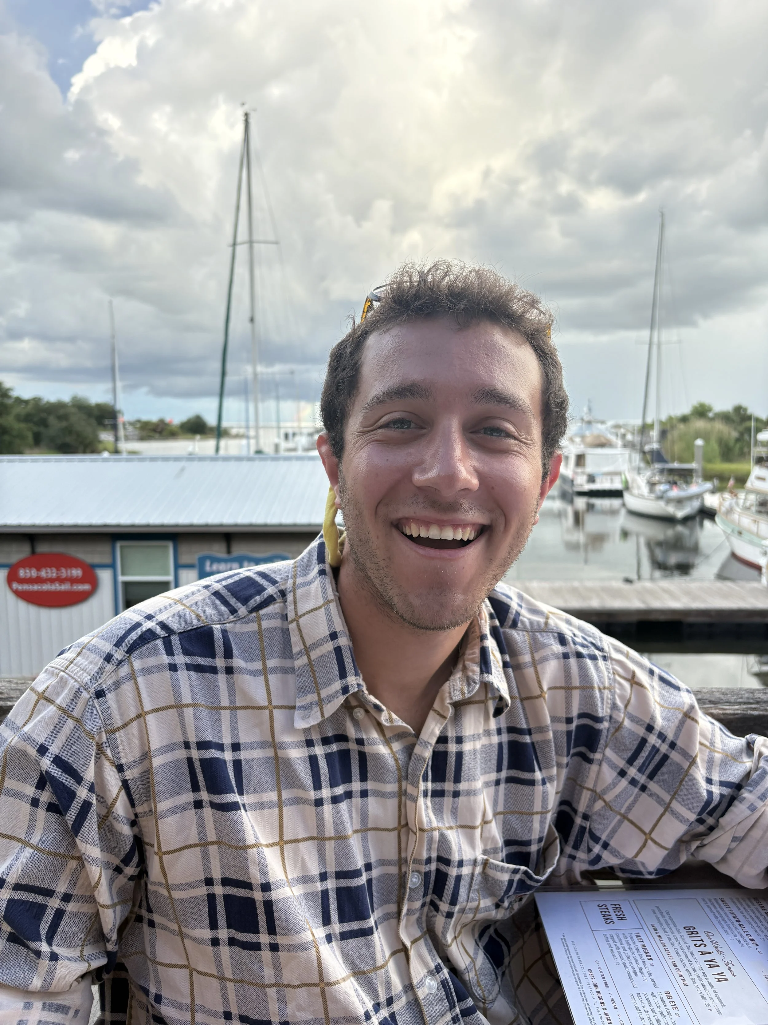 A smiling man with short curly hair wearing a plaid shirt sitting outdoors at a marina with boats and cloudy sky in the background.