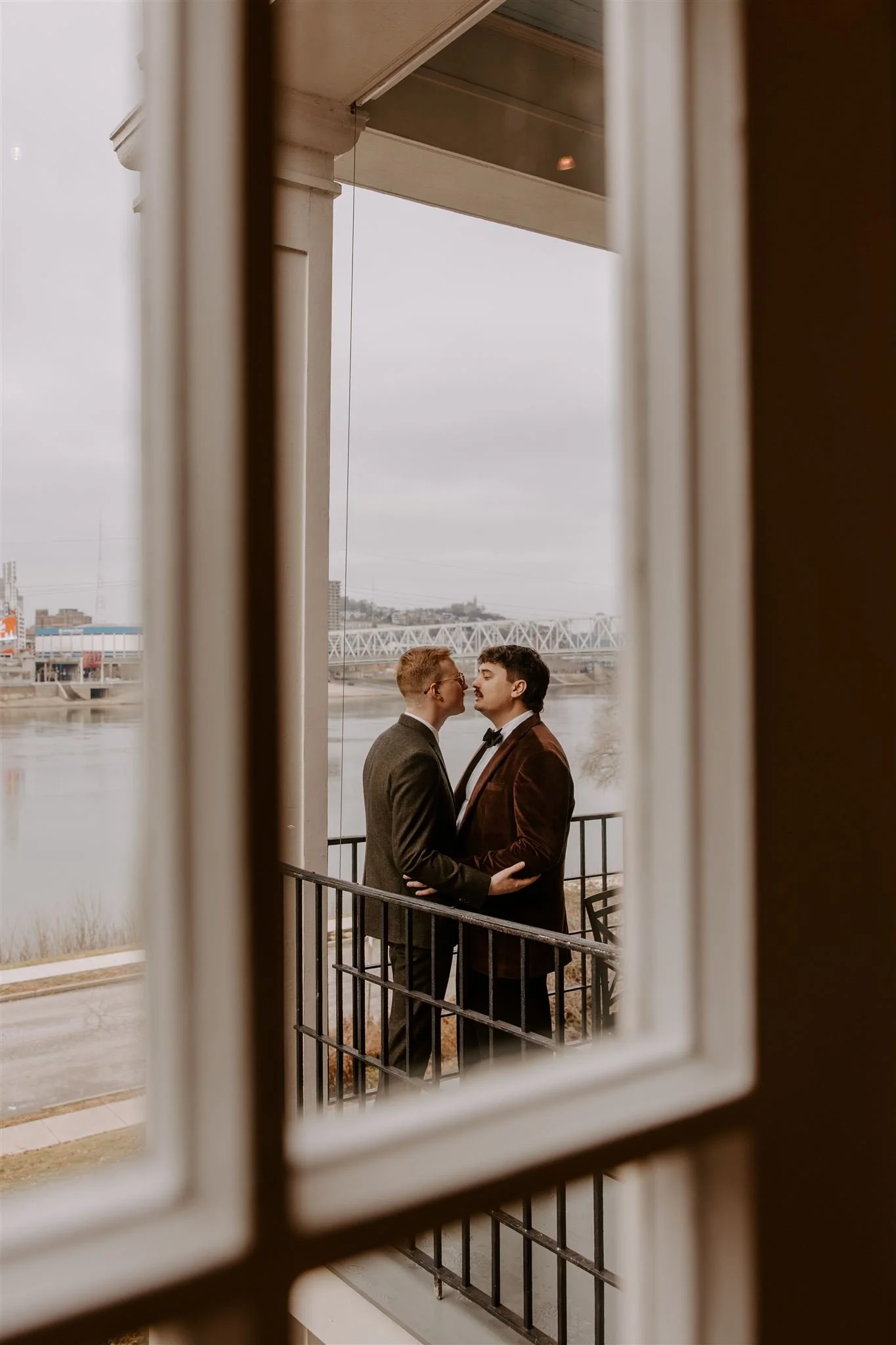 Two men in formal attire standing closely on a balcony, their foreheads touching, viewed through a window with white framing, with an overcast sky and a river in the background.