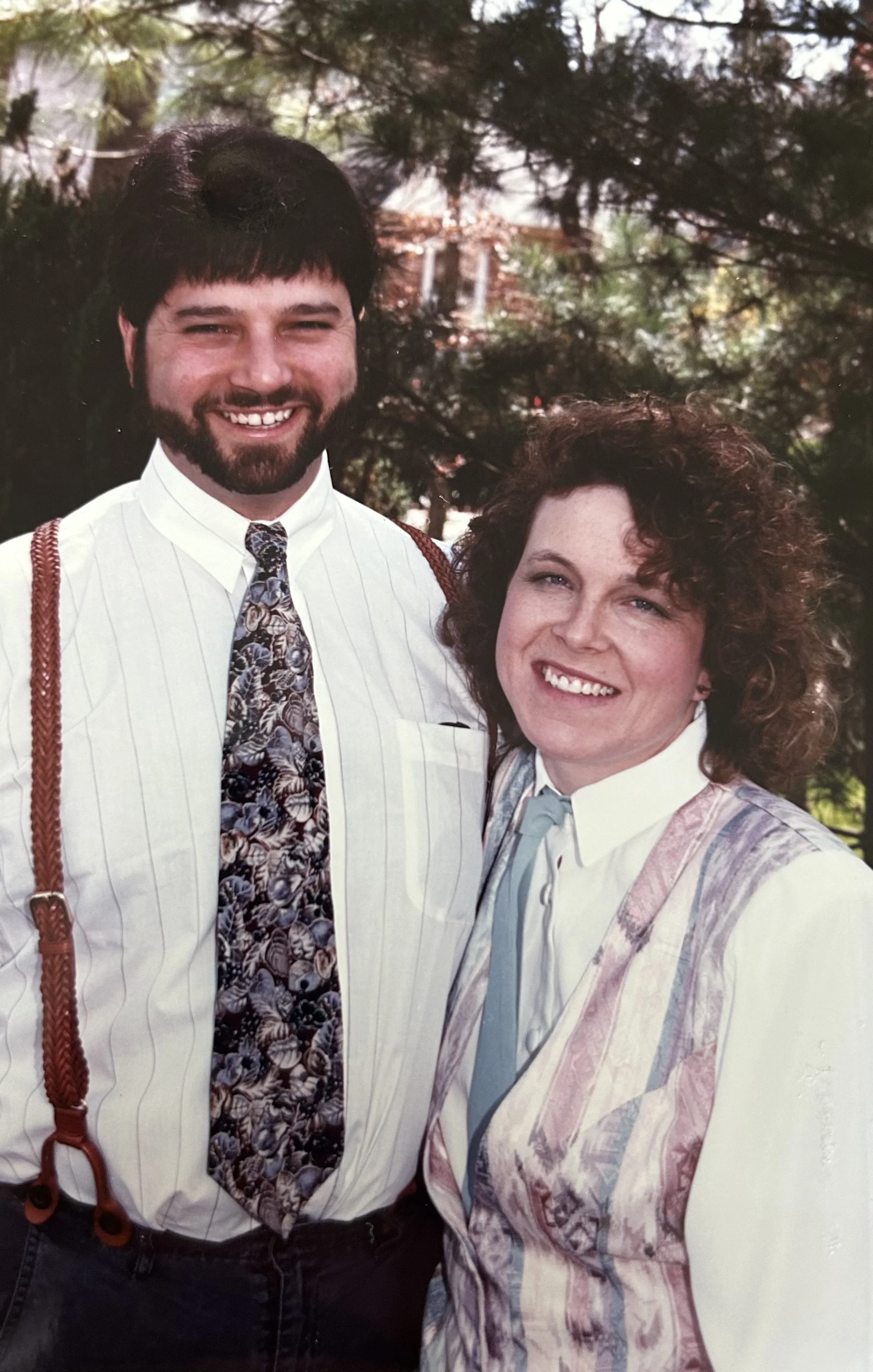 A man with dark hair and a beard smiling wearing a white shirt with a floral tie, suspenders, and a woman smiling with curly hair, wearing a pastel-colored, patterned jacket over a light blue shirt, standing outdoors with green trees and a house in the background.