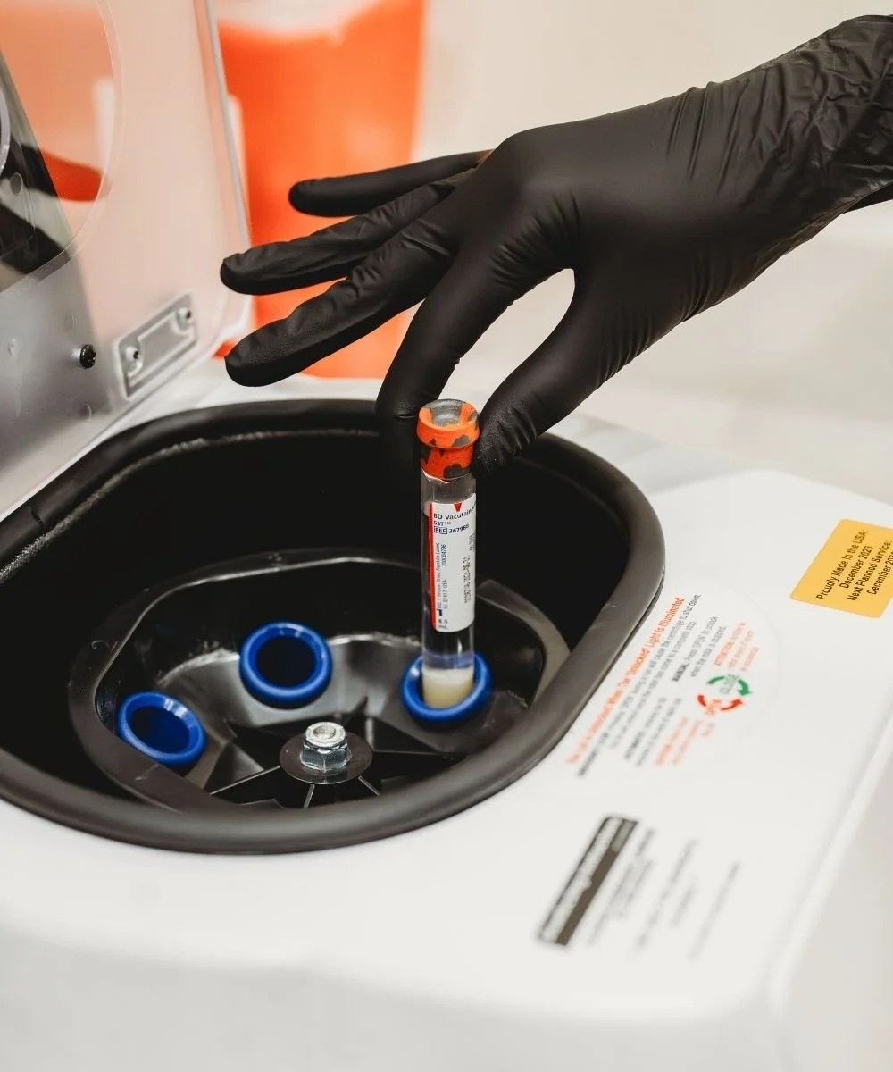 A gloved hand placing a test tube into a medical centrifuge machine.
